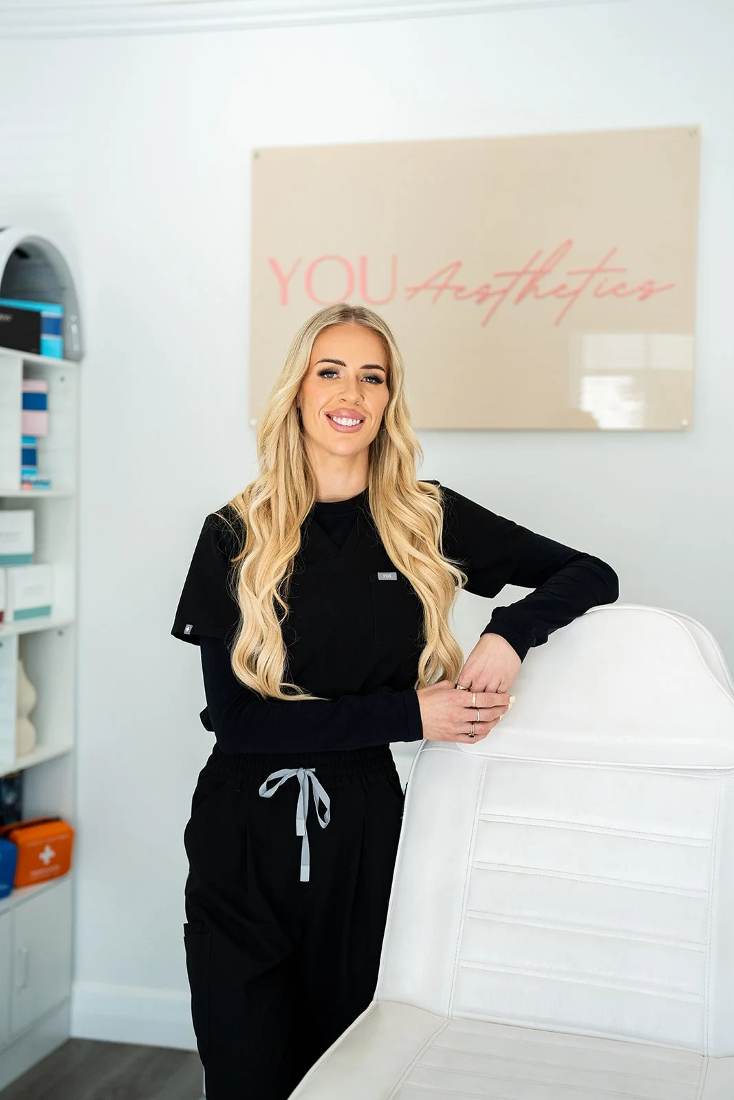 A smiling blonde woman in black medical scrubs leaning on a white examination chair in a clinic with a beige sign that says 'You Aesthetics' in pink cursive lettering on the wall behind her.