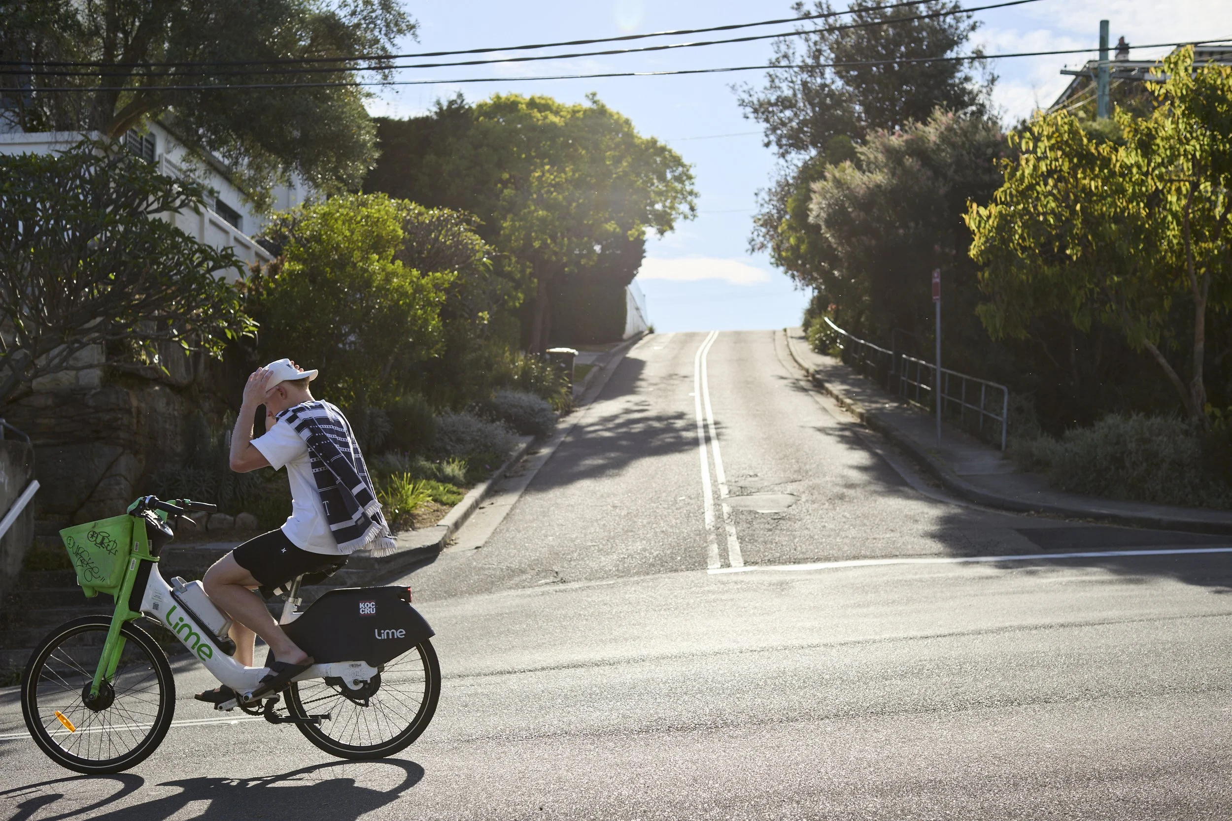 A young boy riding a Lime electric bike on a sunlit street with a hill and greenery in the background.