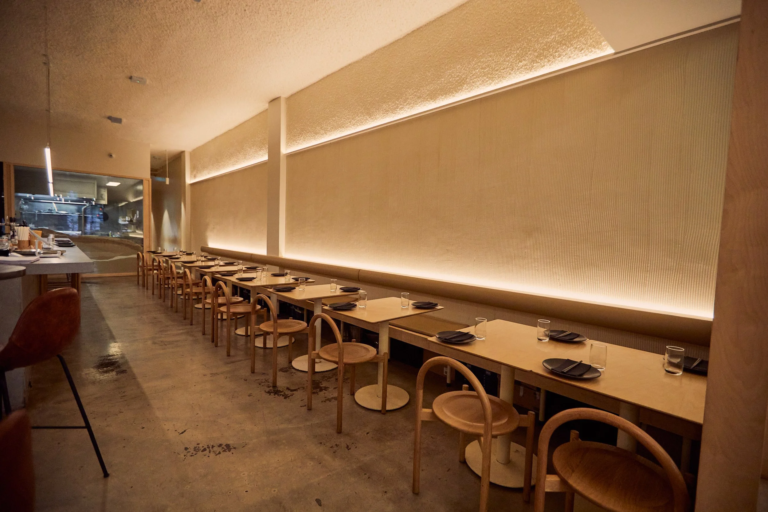 Minimalist restaurant interior with wooden tables and chairs, dim lighting, and an open kitchen view.
