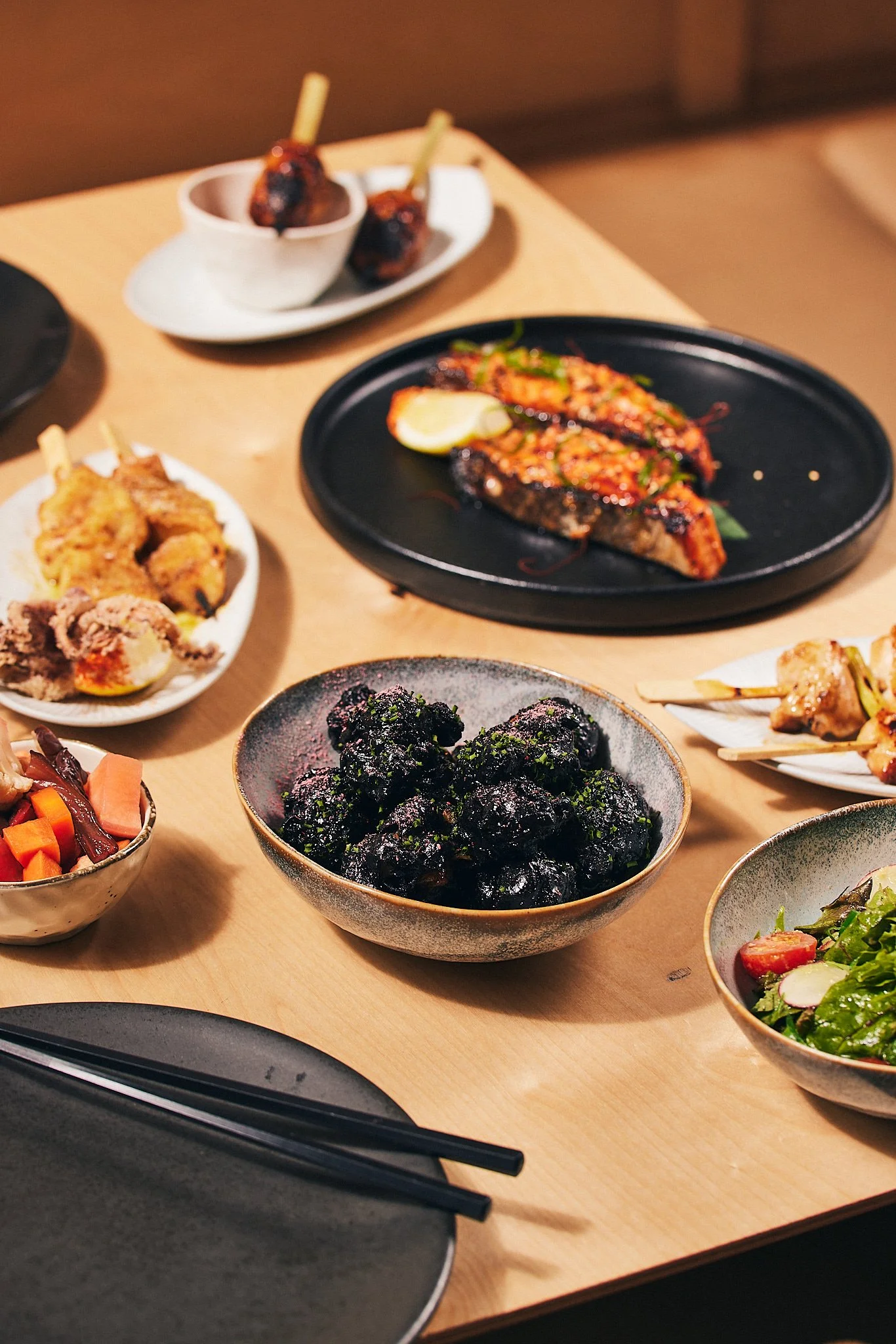 Assorted Japanese dishes on a table, including grilled skewers, vegetables, and dark coated snacks, with chopsticks and plates.