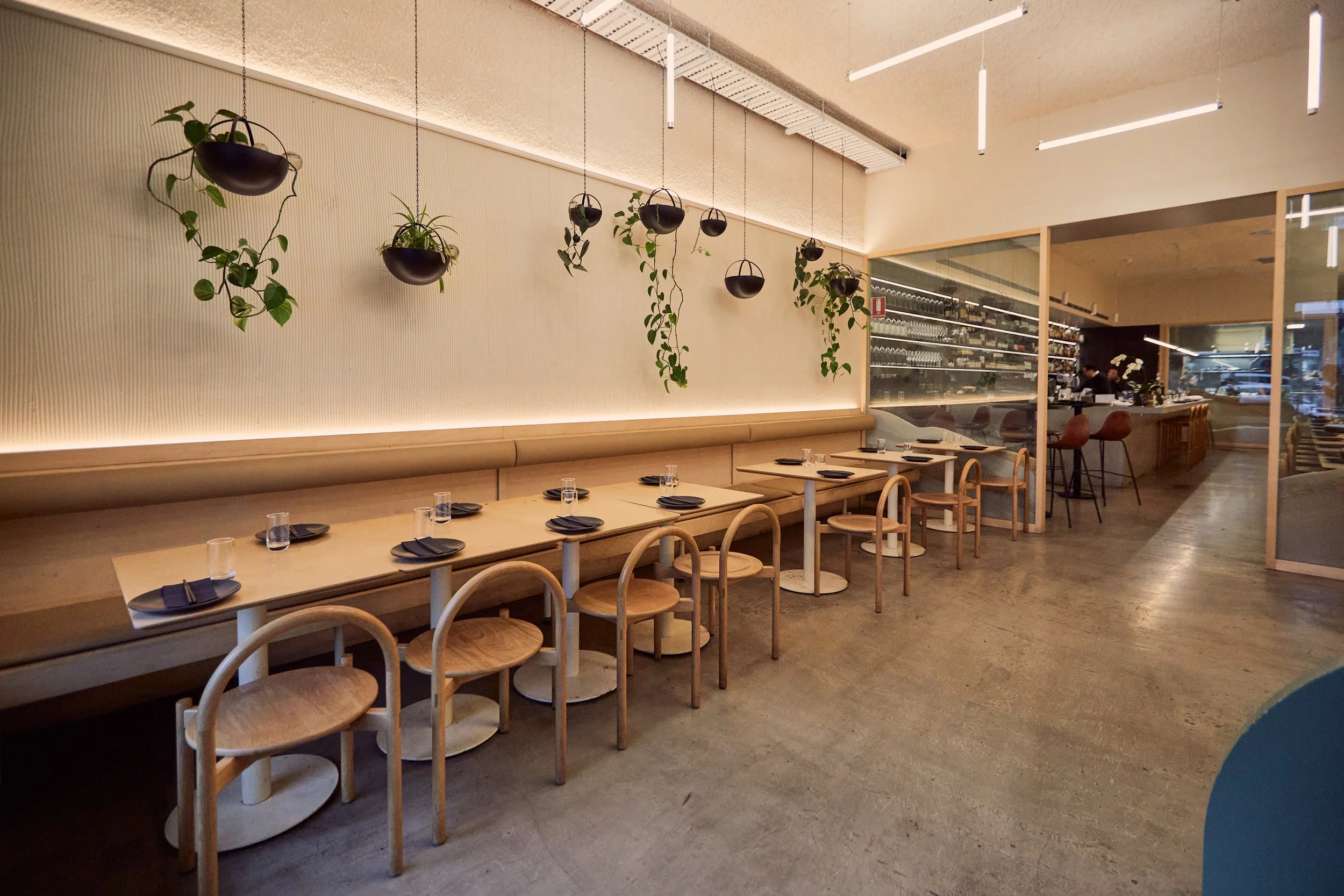 Modern restaurant interior with wooden tables and chairs, hanging plants, minimalist decor, lighting fixtures, and a visible bar area in the background.