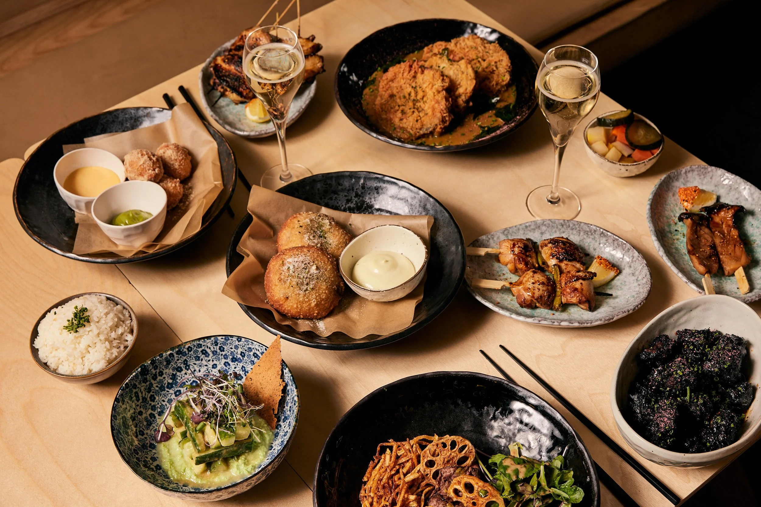 Assorted Japanese dishes on a table, featuring skewers, fried croquettes, rice, vegetable dishes, and two glasses of bubbly wine.