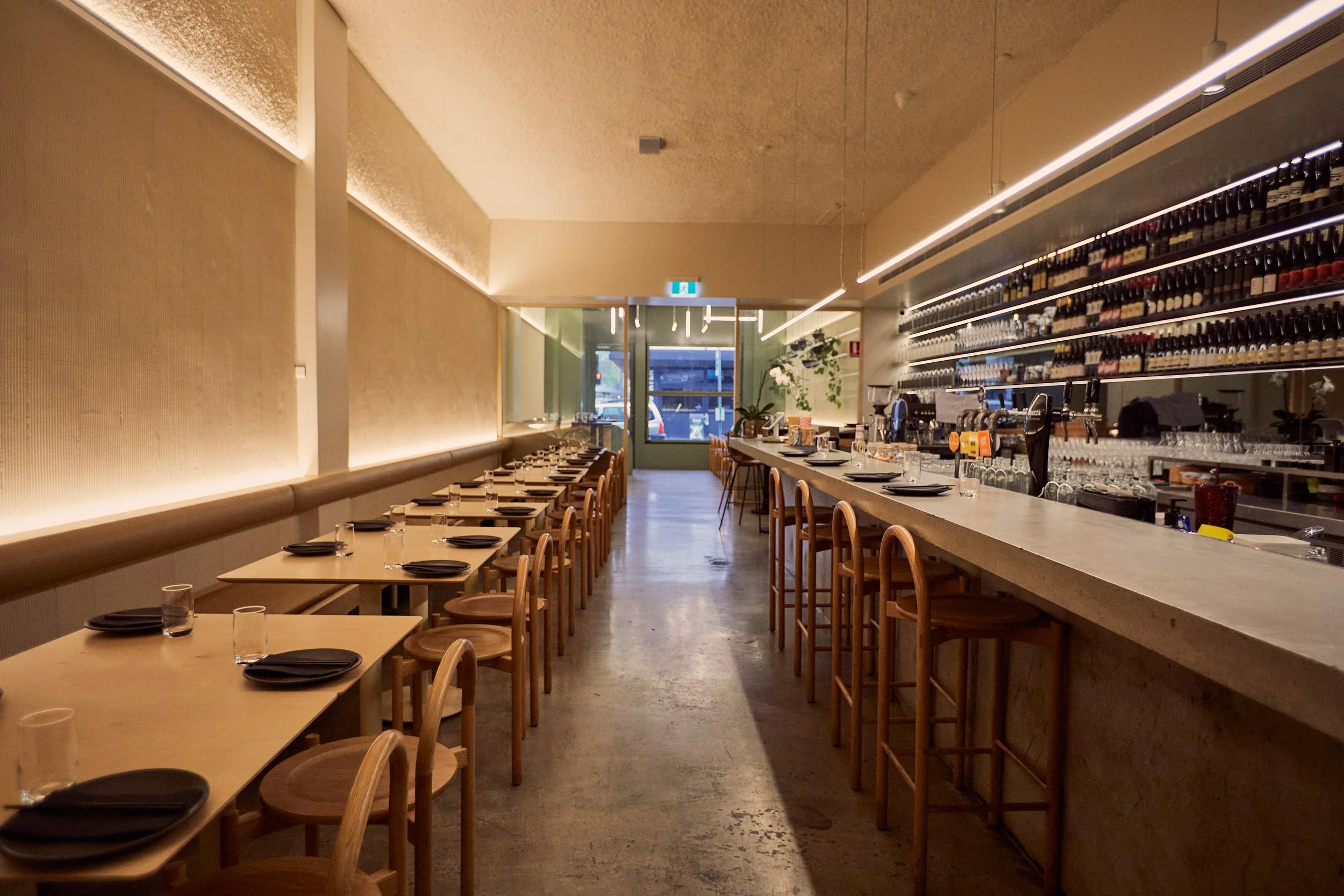 Modern restaurant interior with wooden tables and chairs, bar counter, wine bottles on shelves, and soft ambient lighting.