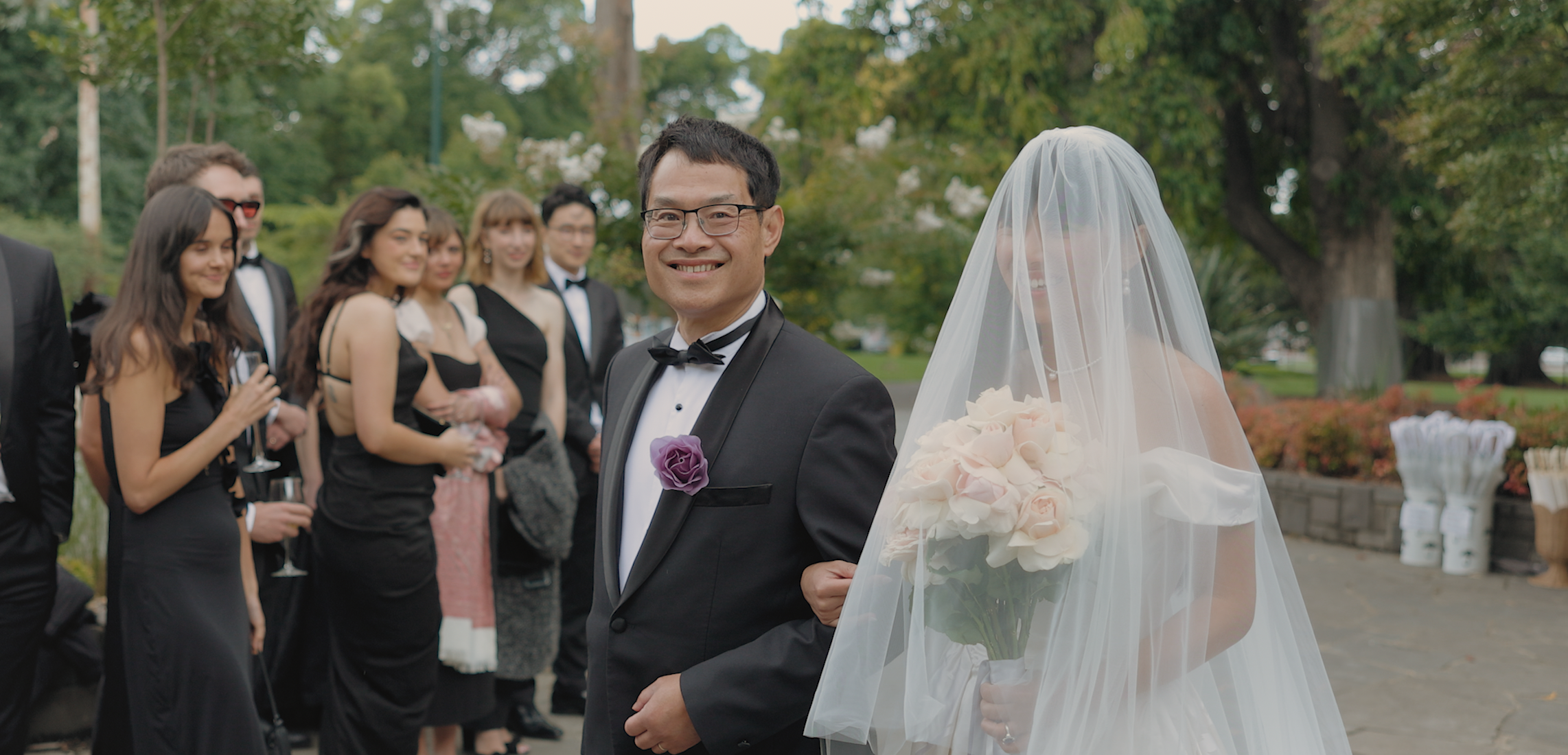 A happy groom standing outdoors with a bouquet of pink roses, smiling at the camera, as a group of wedding guests in formal attire gather behind him. The bride, with her face partially obscured by a veil, stands next to him holding a similar bouquet. The scene is set in a lush, green park.
