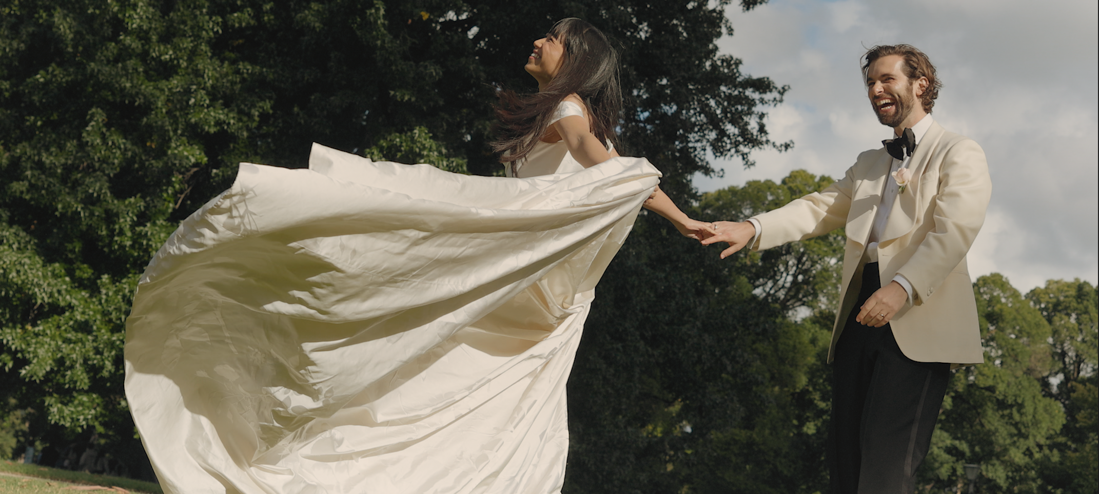 A bride and groom holding hands and laughing outdoors, with trees and a cloudy sky in the background.