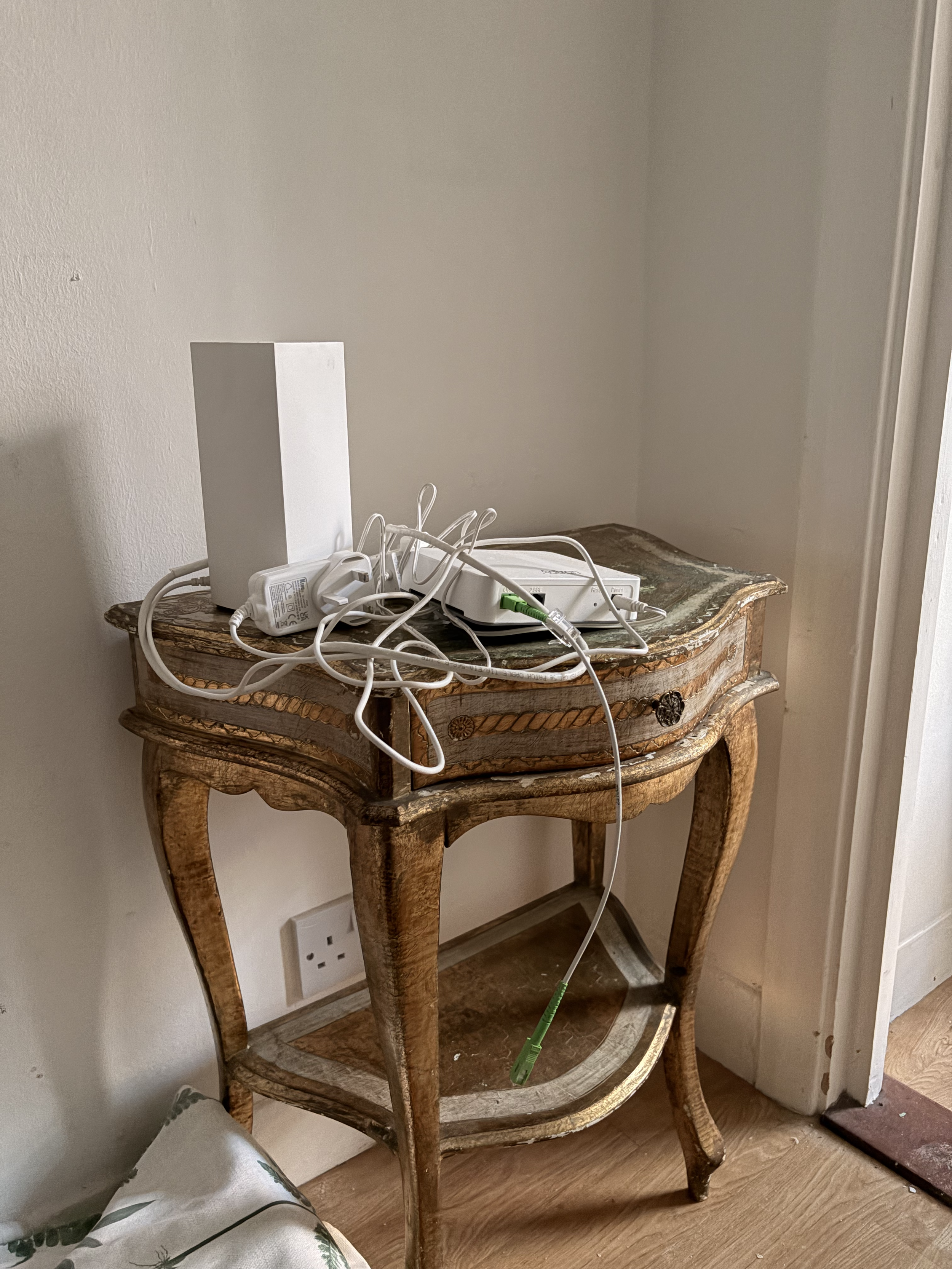 An antique wooden side table with a clutter of electronic cords, a power strip, and a white Wi-Fi router on top. A white power outlet with a plug is visible on the wall behind the table. The table is placed next to a white wall and window with a white curtain.