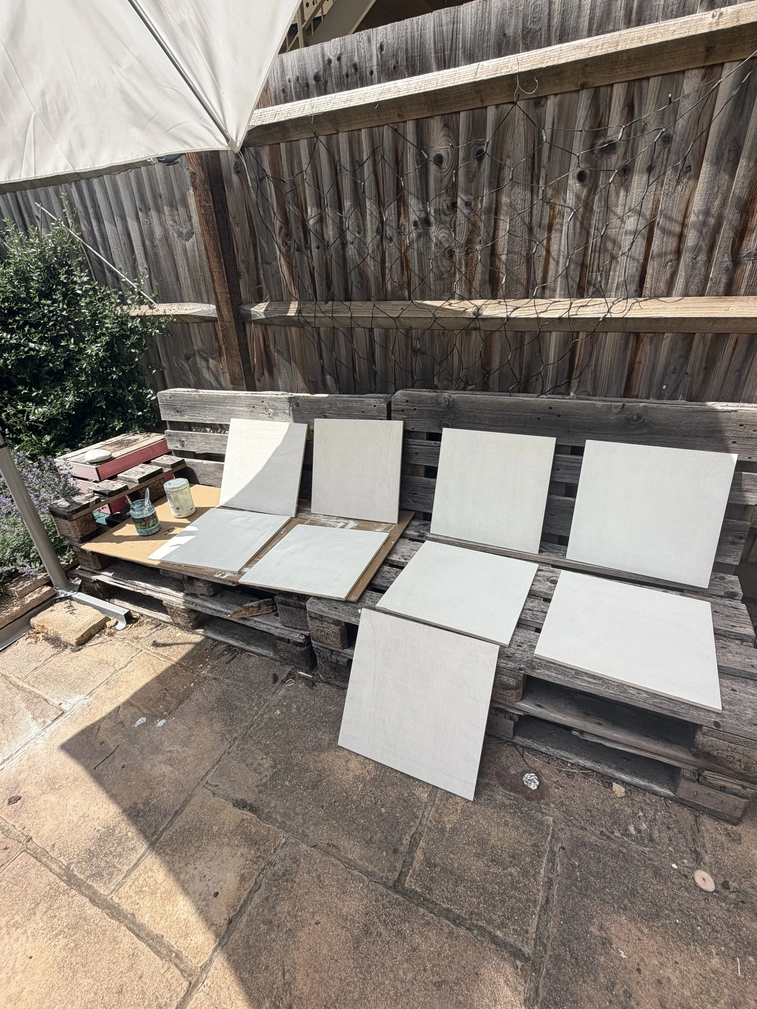 Outdoor scene with a weathered wooden fence, partially covered by a white umbrella. Several white painted tiles are leaning against a wooden bench, with cans of paint and a small brick structure nearby. The ground is paved with stone tiles.