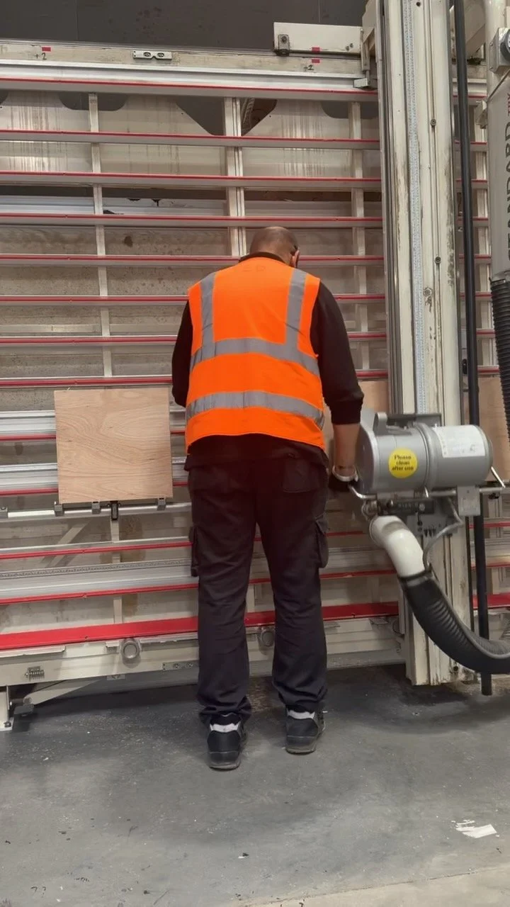 A worker in an orange safety vest uses a power tool on a large industrial machine inside a warehouse or factory.