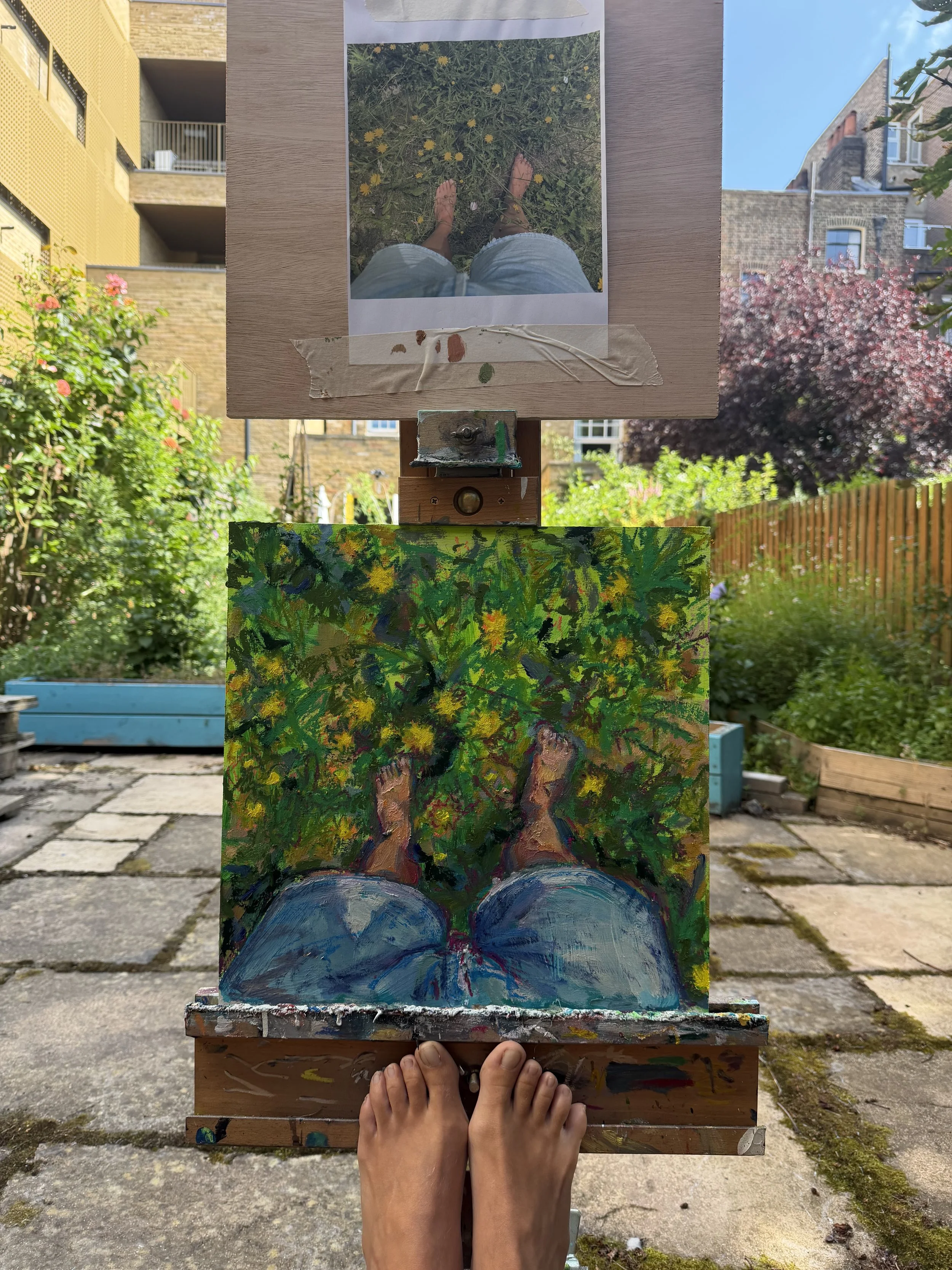 A person viewing their painted artwork outdoors on a wooden easel with urban buildings and greenery in the background.