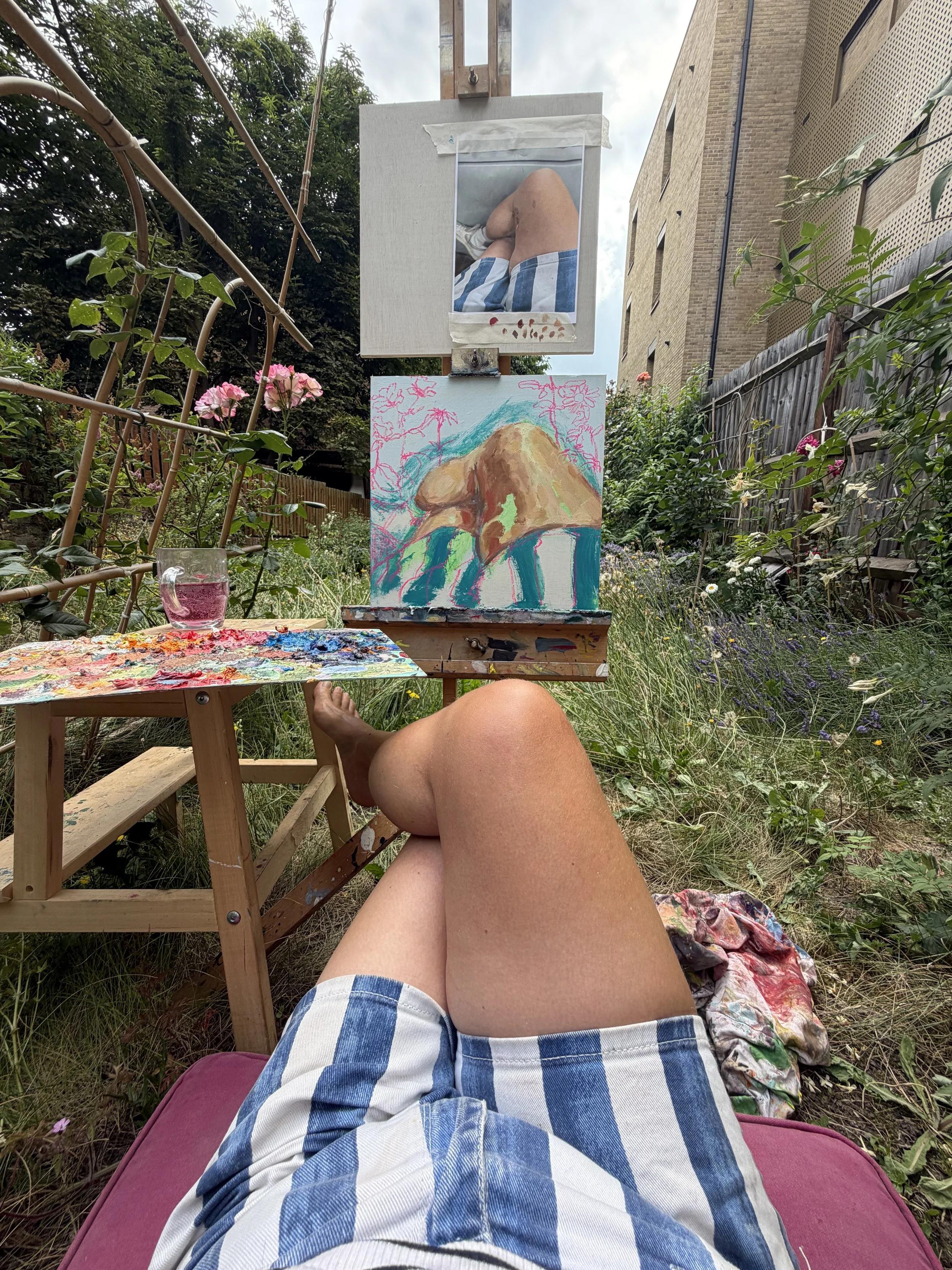 A person in a blue and white checkered shirt is sitting outdoors on a pink cushion, with painted artwork on an easel in front. The artwork depicts colorful abstract figures, and the person is taking a photo of their own legs and the painting. The scene is in a garden with plants, flowers, and a wooden fence around.