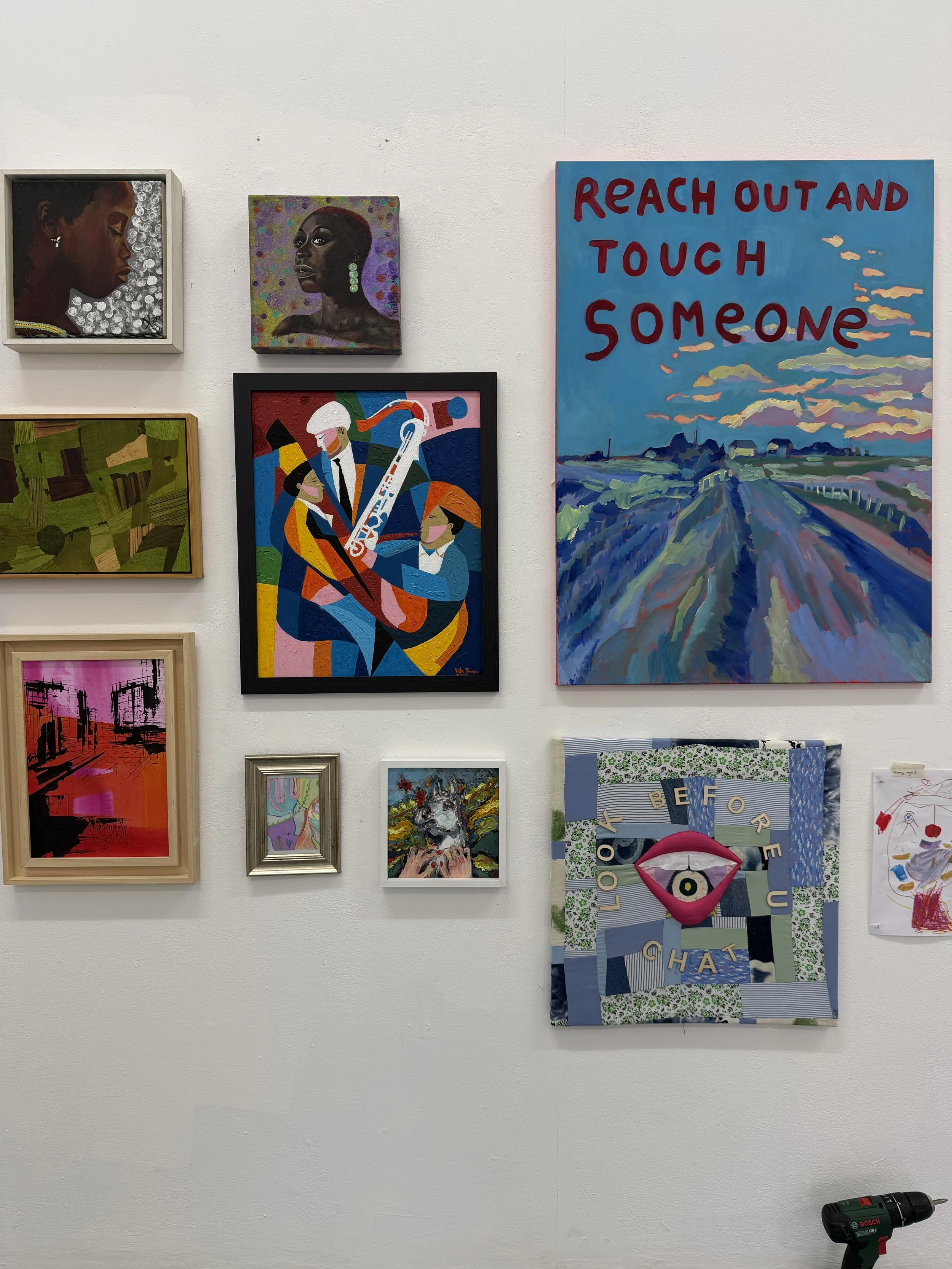 A collection of small and large colorful paintings on a white wall, including portraits, abstract scenes, and a large blue painting with the words "Reach Out and Touch Someone." There is also a cordless power drill on the bottom right corner of the i