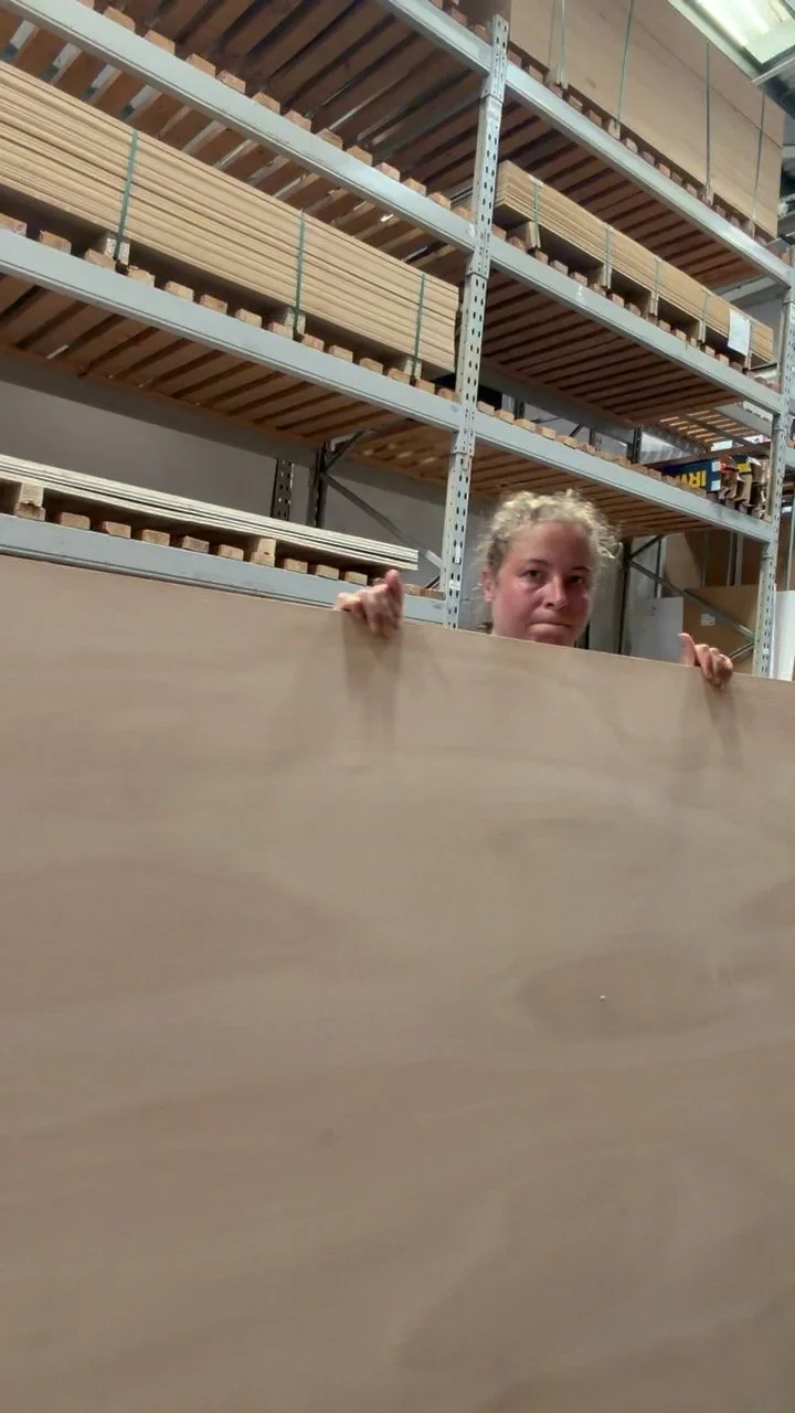 A young girl peeks over a large wooden board in a hardware or woodworking store, with shelves of wood and other supplies in the background.