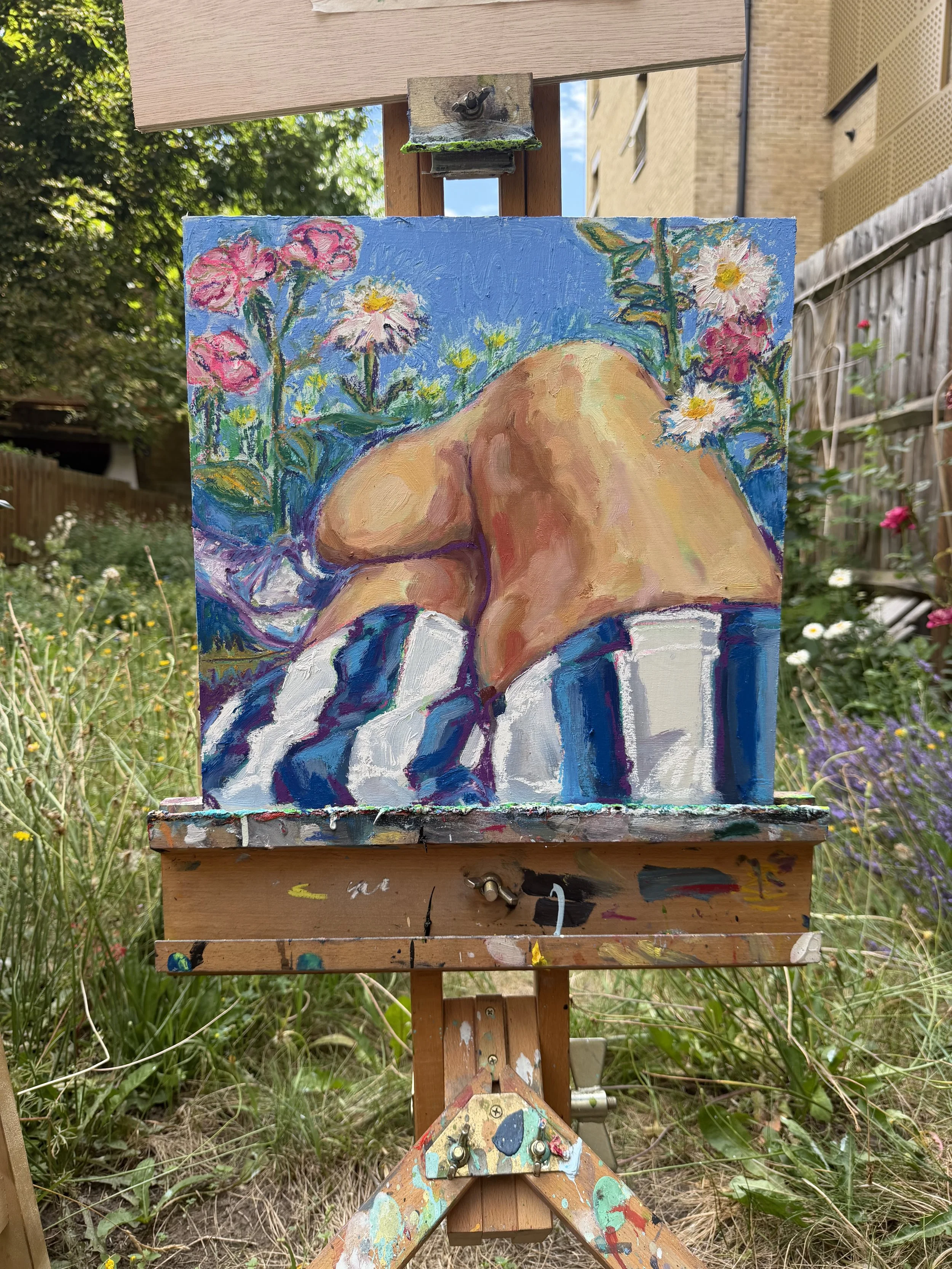 Painting of a nude figure with flowers and a blue background set outdoors on an easel.