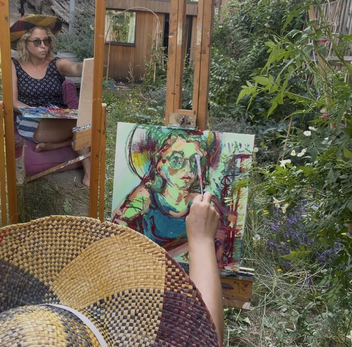 A person painting a colorful portrait outdoors, with a woman sitting inside a woodworking or gardening shed visible in the mirror reflection.