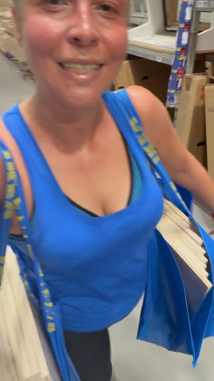 A woman with blonde hair smiling while shopping at a home improvement store, wearing a blue tank top and carrying a blue shopping bag.
