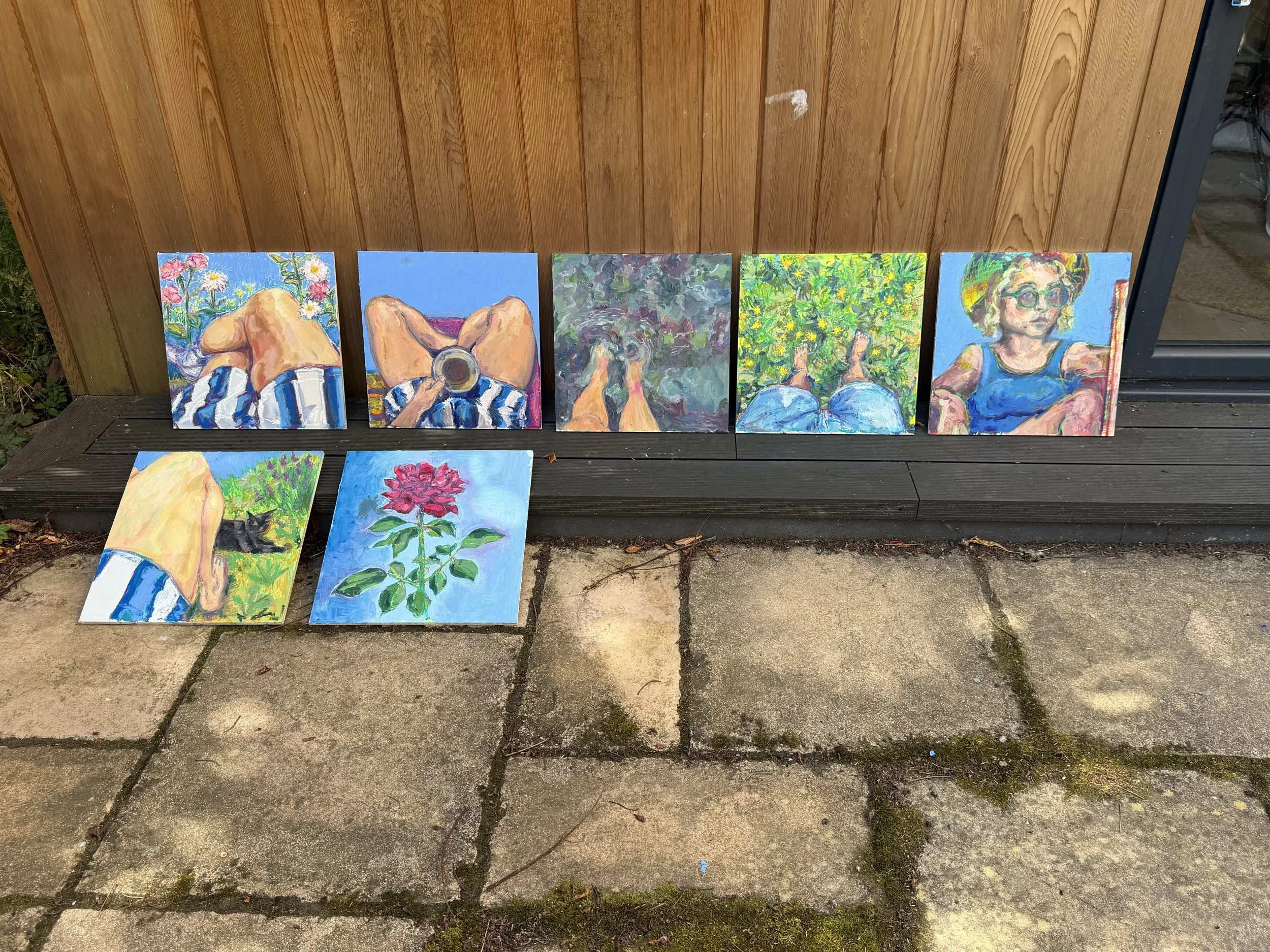 A collection of colorful impressionist-style paintings of various scenes, including a woman taking a photograph, a dog, plants, and a girl with glasses, displayed on a wooden outdoor wall and resting on the ground.