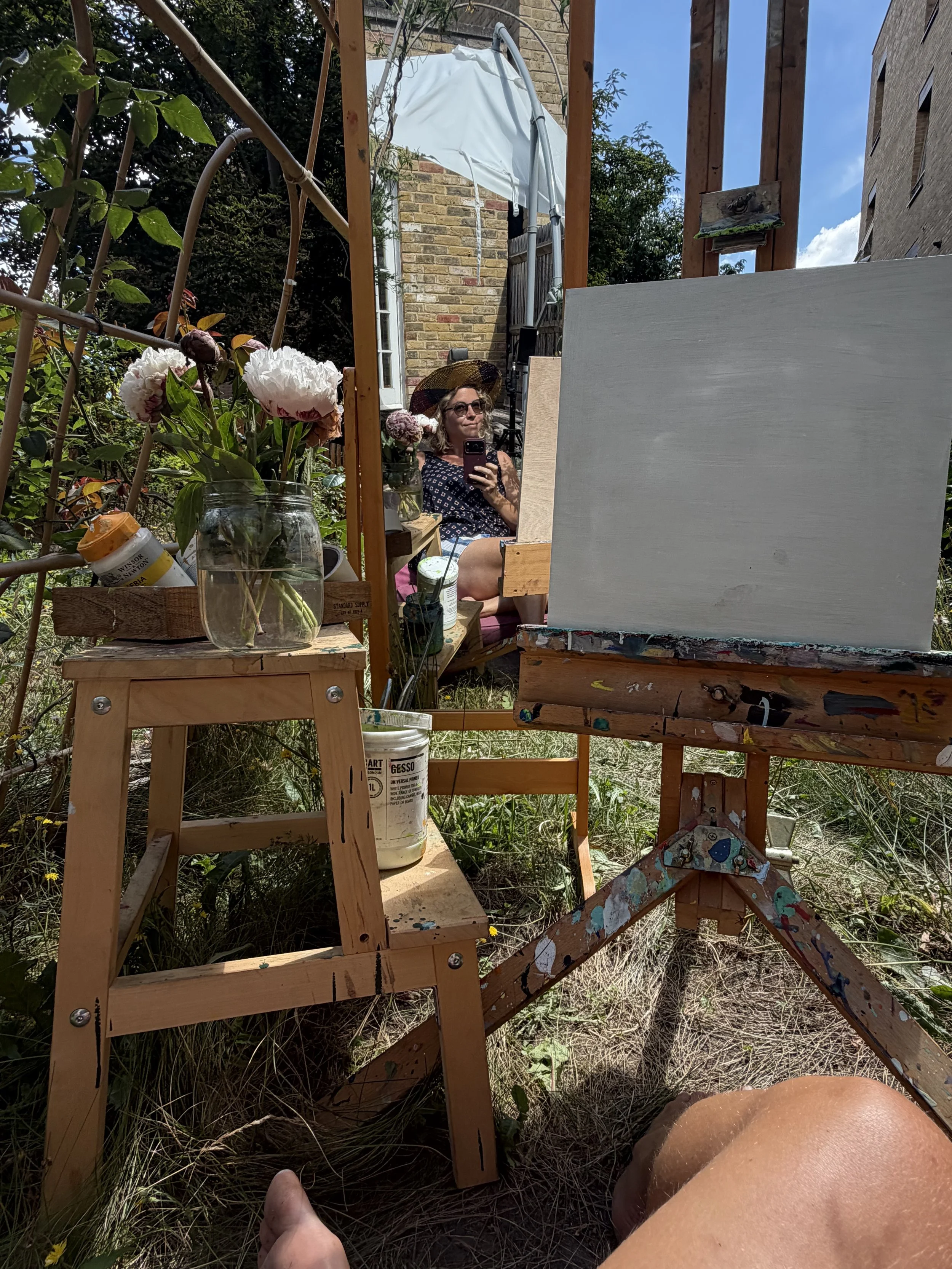 A woman sitting outdoors in front of an easel with an empty canvas, wearing a wide-brimmed hat and sunglasses, surrounded by plants and flowers, taking a selfie with her phone.