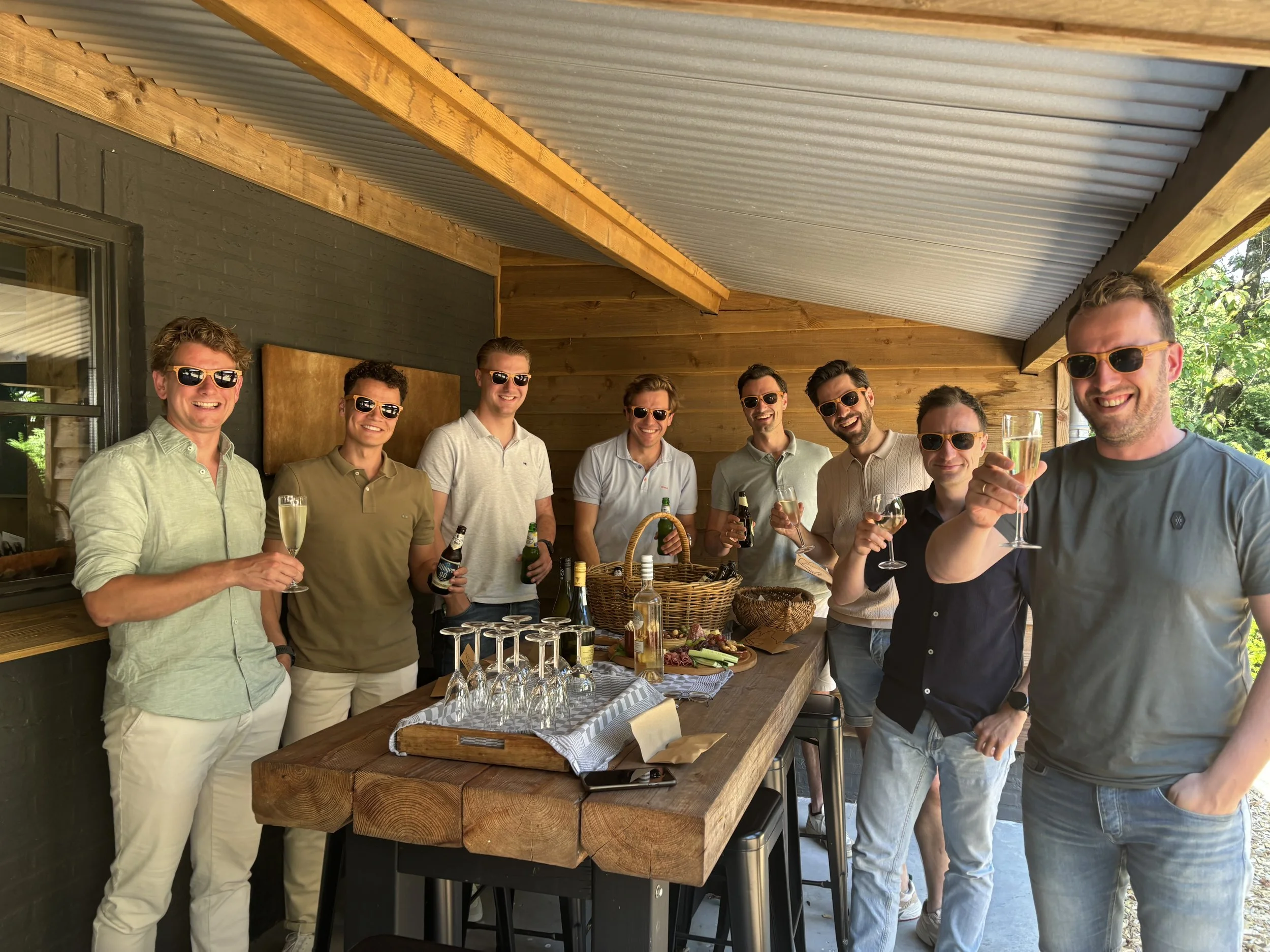 Een groep mensen die samen proosten met champagne en bier, staand achter een tafel met drank en eten op een veranda.