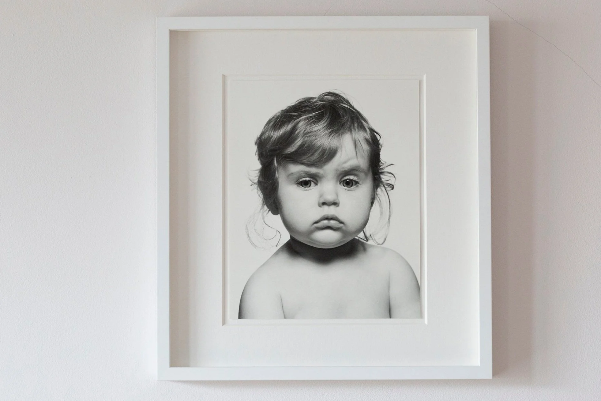 Black and white photograph of a young child with large expressive eyes and tousled hair, portrayed in a framed portrait on a white wall.