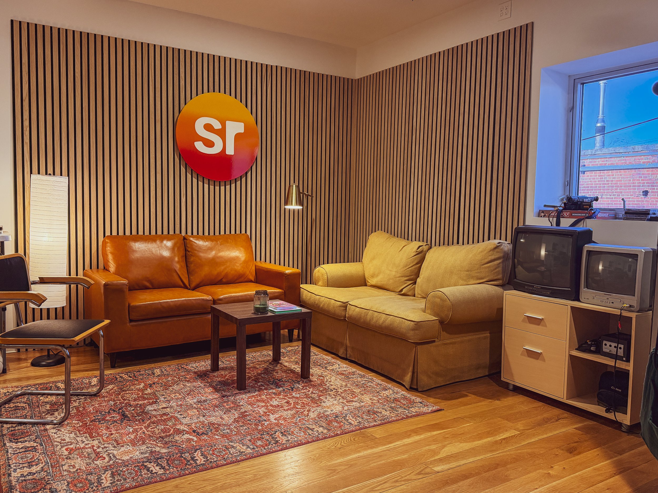 Living room with two sofas (one tan and one yellow), a wooden coffee table, an area rug, a TV stand with two vintage TVs, a window showing a brick building outside, and a wooden wall with a red and orange logo featuring the initials 'sr' on it.