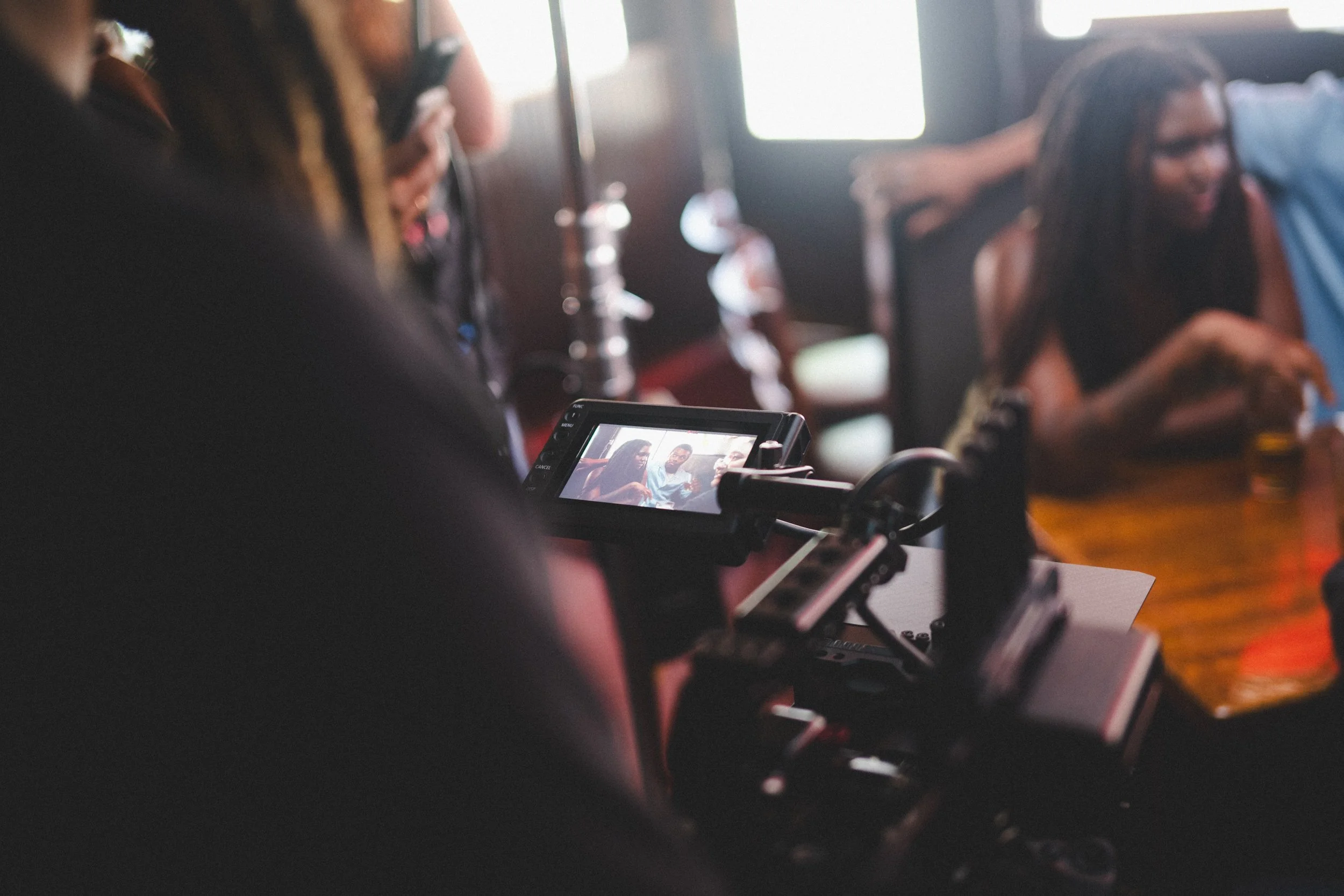 A camera on a tripod is capturing a scene of two women sitting at a wooden table in a dimly lit restaurant or pub. One woman has long braids, and it appears she is talking or eating.