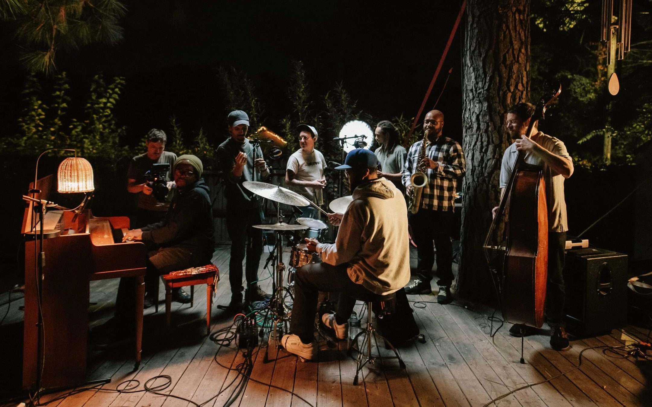 A group of musicians performing outdoors at night, including a pianist, drummer, upright bassist, and wind instrument players, surrounded by trees and warm lighting.