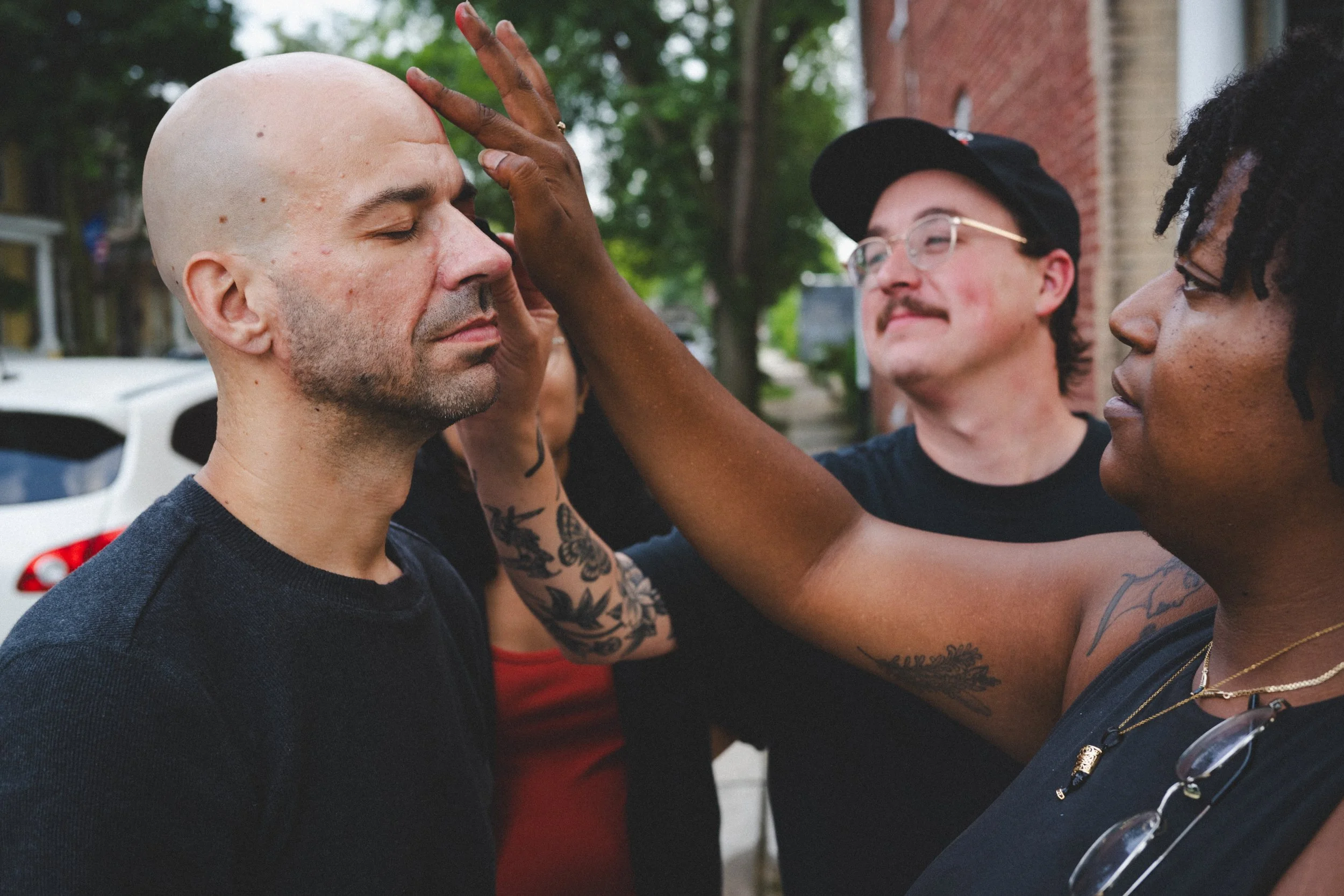 A woman with dark skin, tattoos, and glasses holds a man's face gently while some people watch, on a city street during daytime.