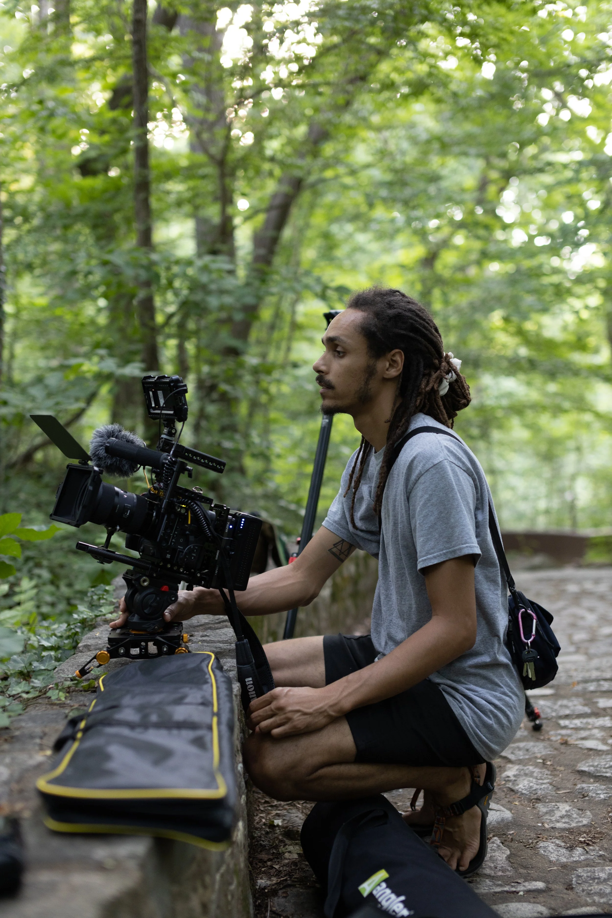 A man with dreadlocks is sitting on a stone wall in a wooded area, operating a professional video camera with various accessories.