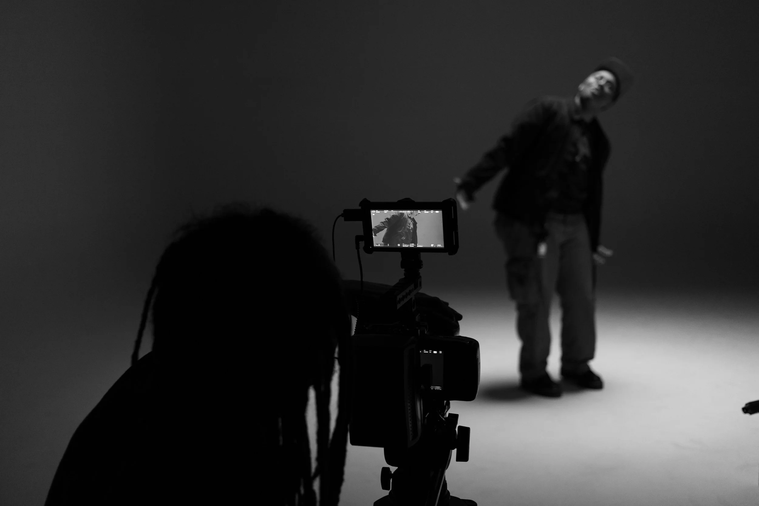 Black and white photo of a person filming a man posing in a photography studio with a plain background.