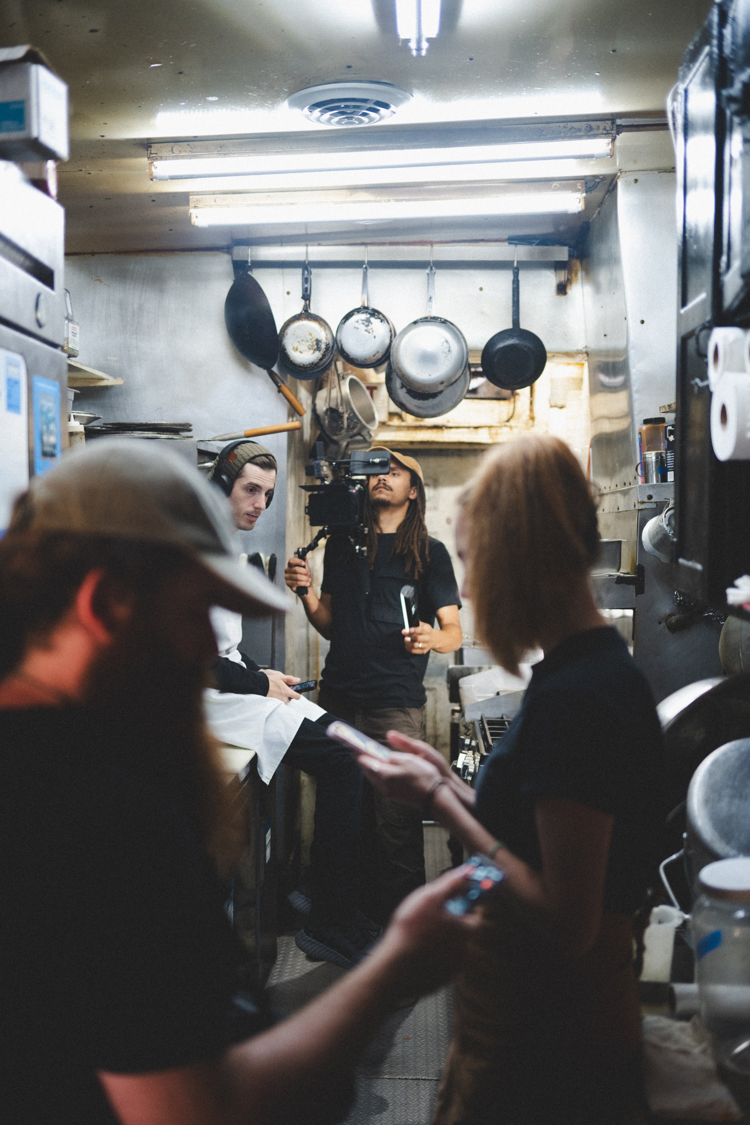 People in a small commercial kitchen with pots hanging on the wall, some individuals using their phones, and a person operating a camera.