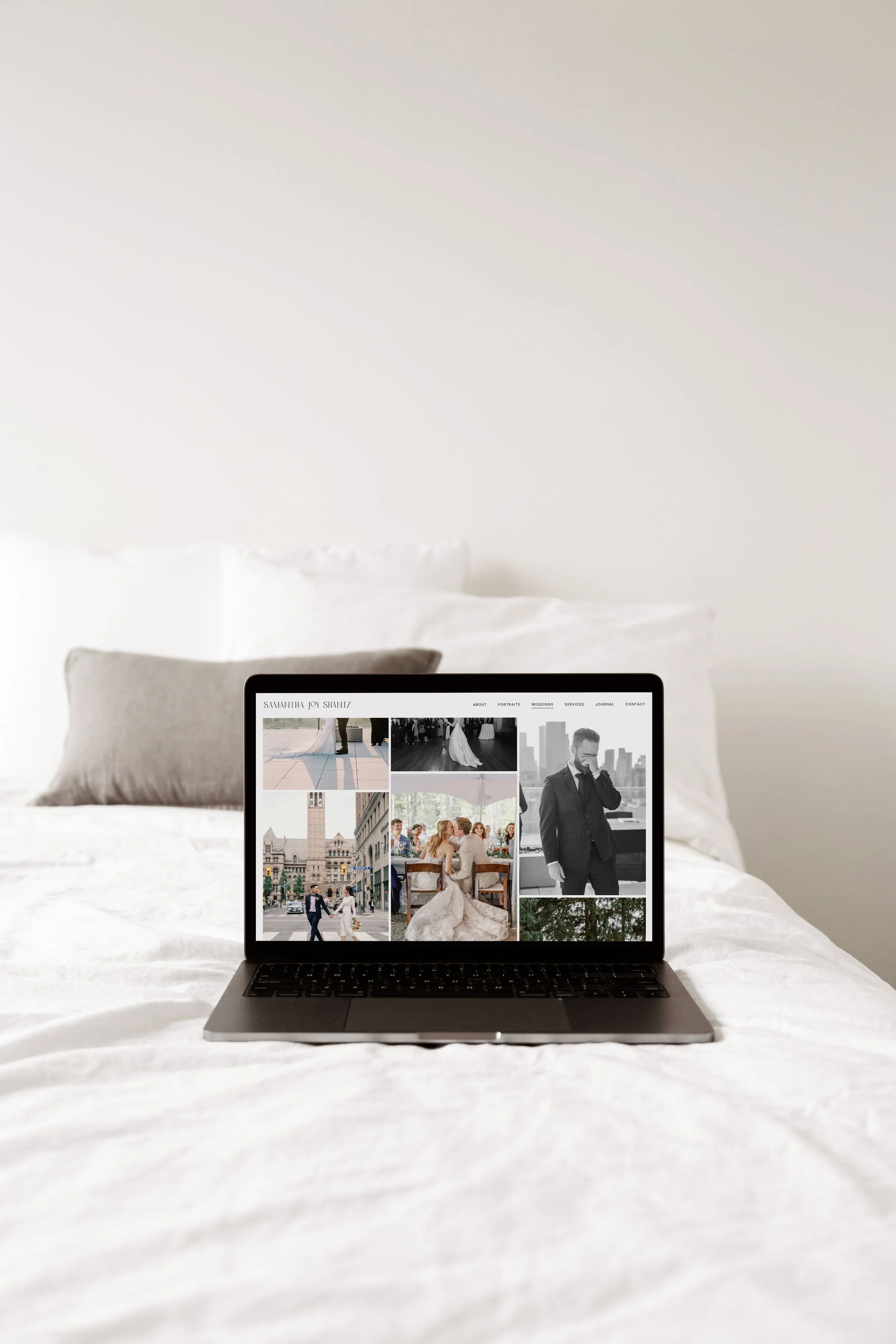 Open laptop displaying a wedding photography website, placed on a white bed with pillows in the background.