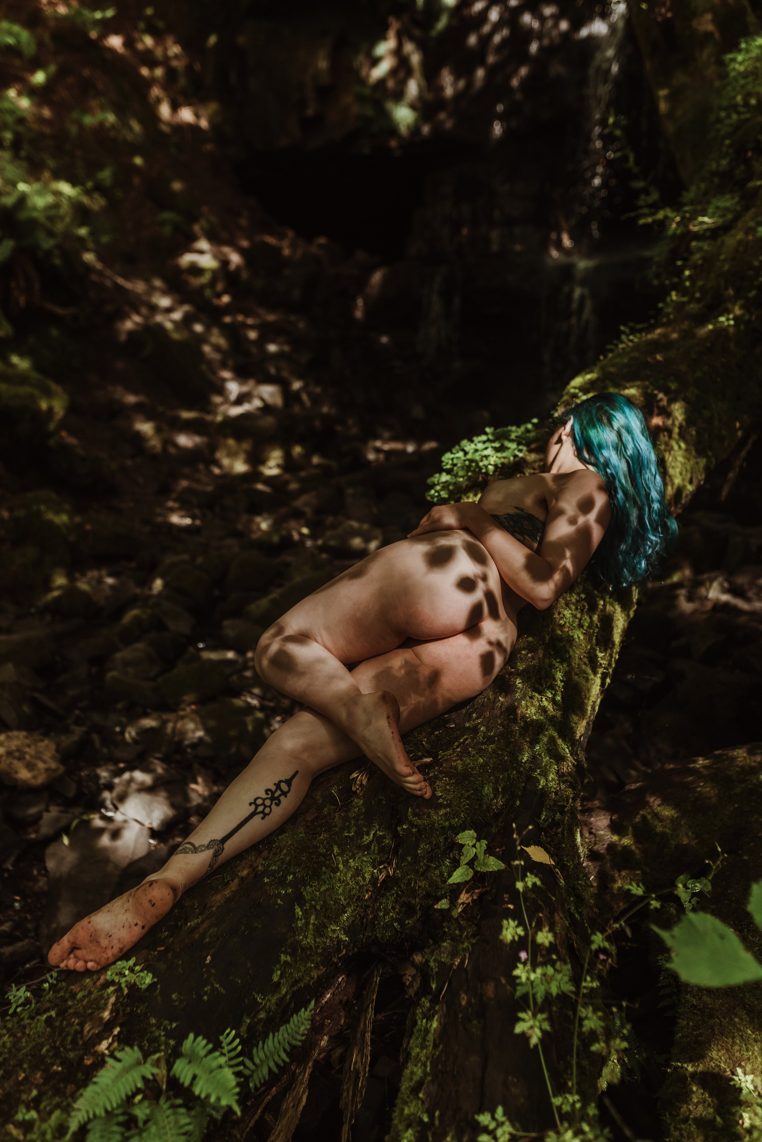 Person with blue hair lying on a moss-covered log in a forest, with dappled sunlight and a tattoo on their leg.