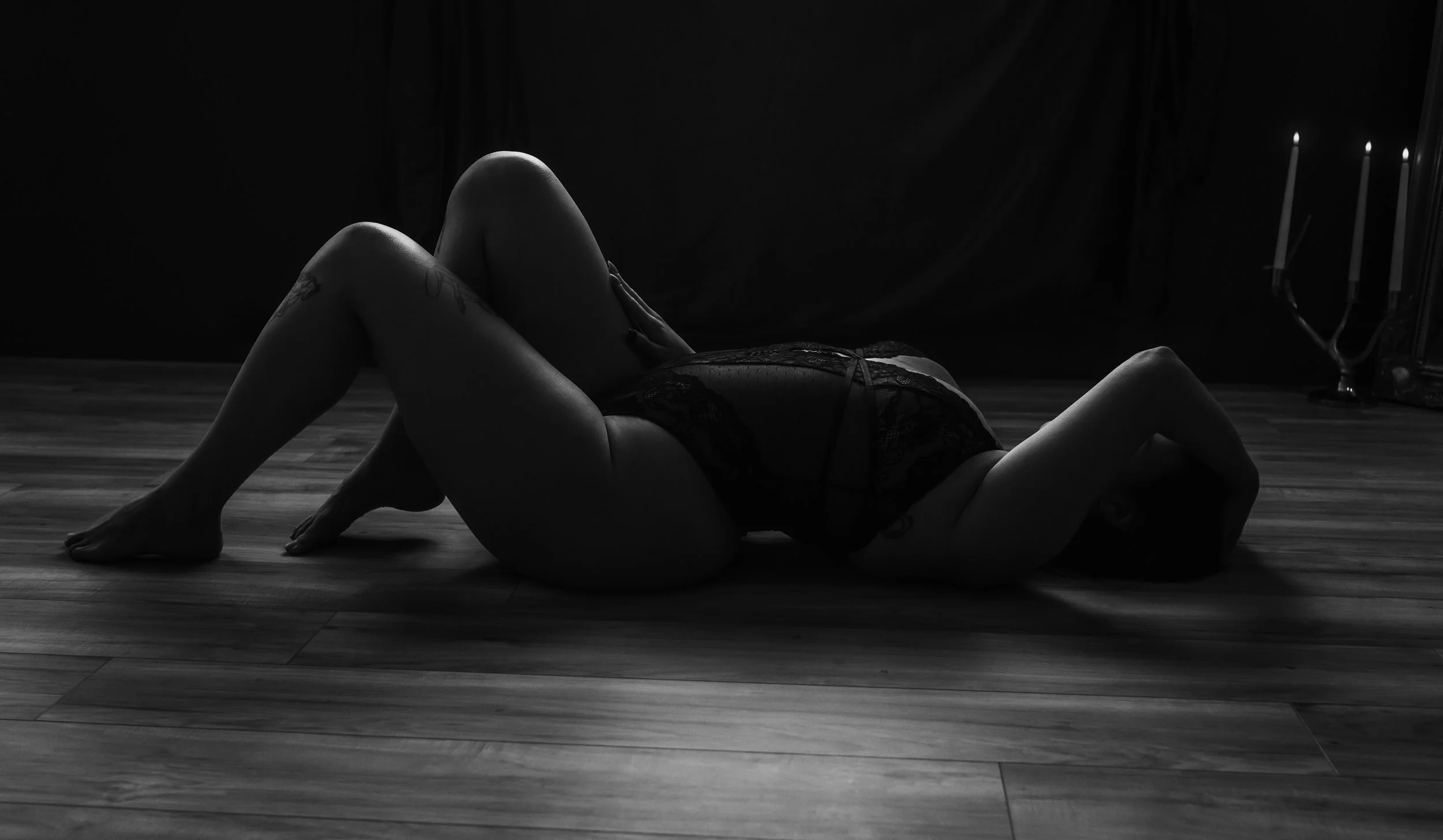 Artistic black and white boudoir photograph taken at Marieke Captures studio Glasgow — woman lying on studio floor in black lace lingerie with candlelight in background, moody and intimate