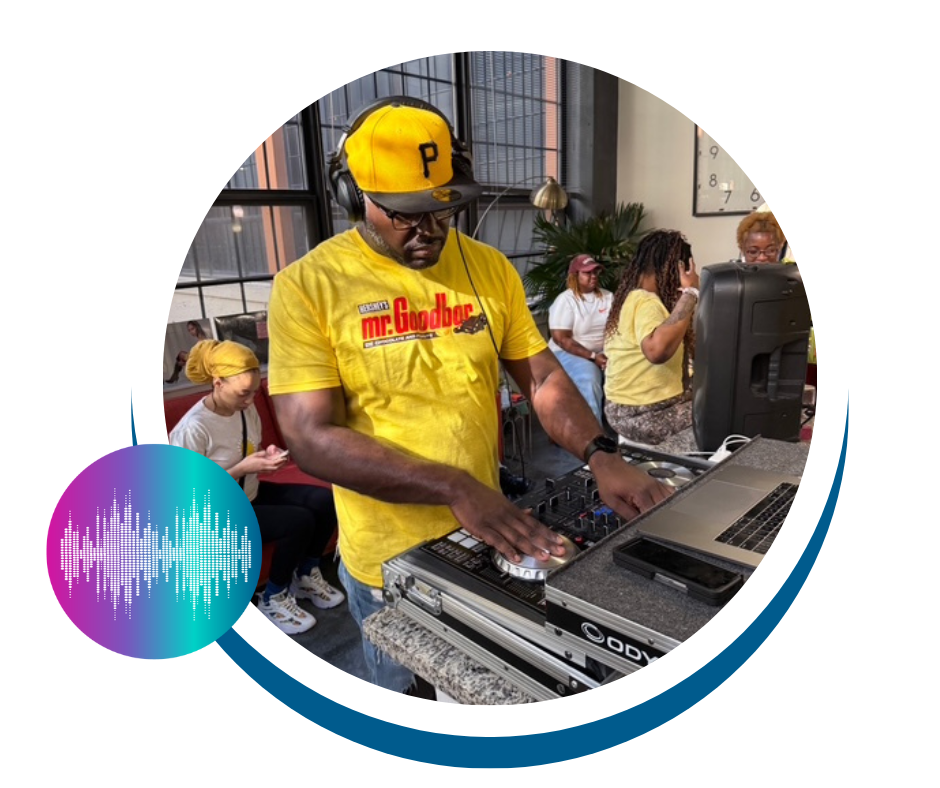 DJ wearing yellow 'mr. Goodbar' t-shirt and a yellow cap with a black 'P' logo, mixing music at a DJ booth in a busy indoor setting with several people in the background.