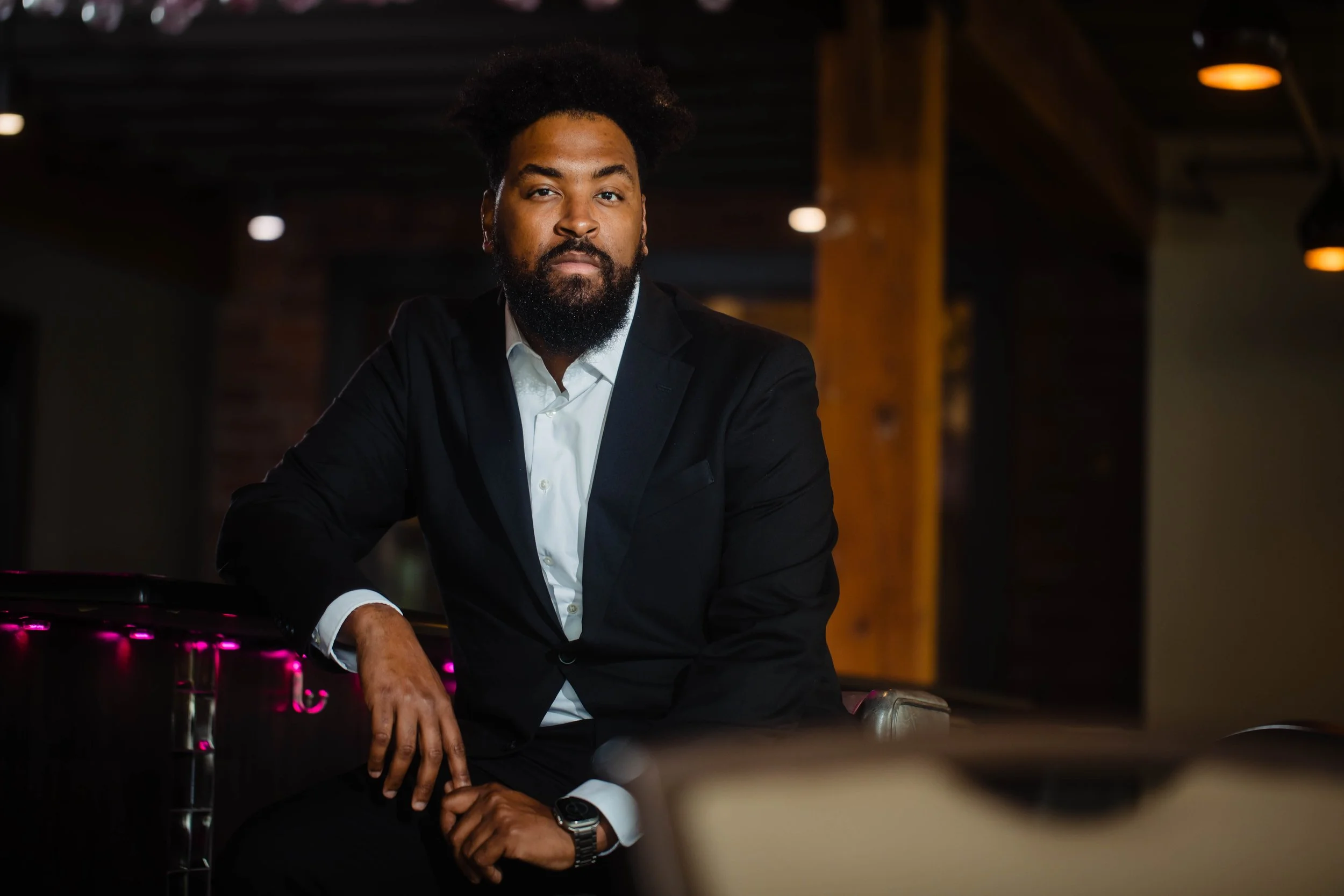 A man wearing a black suit and white shirt sitting indoors at a table with a serious expression.