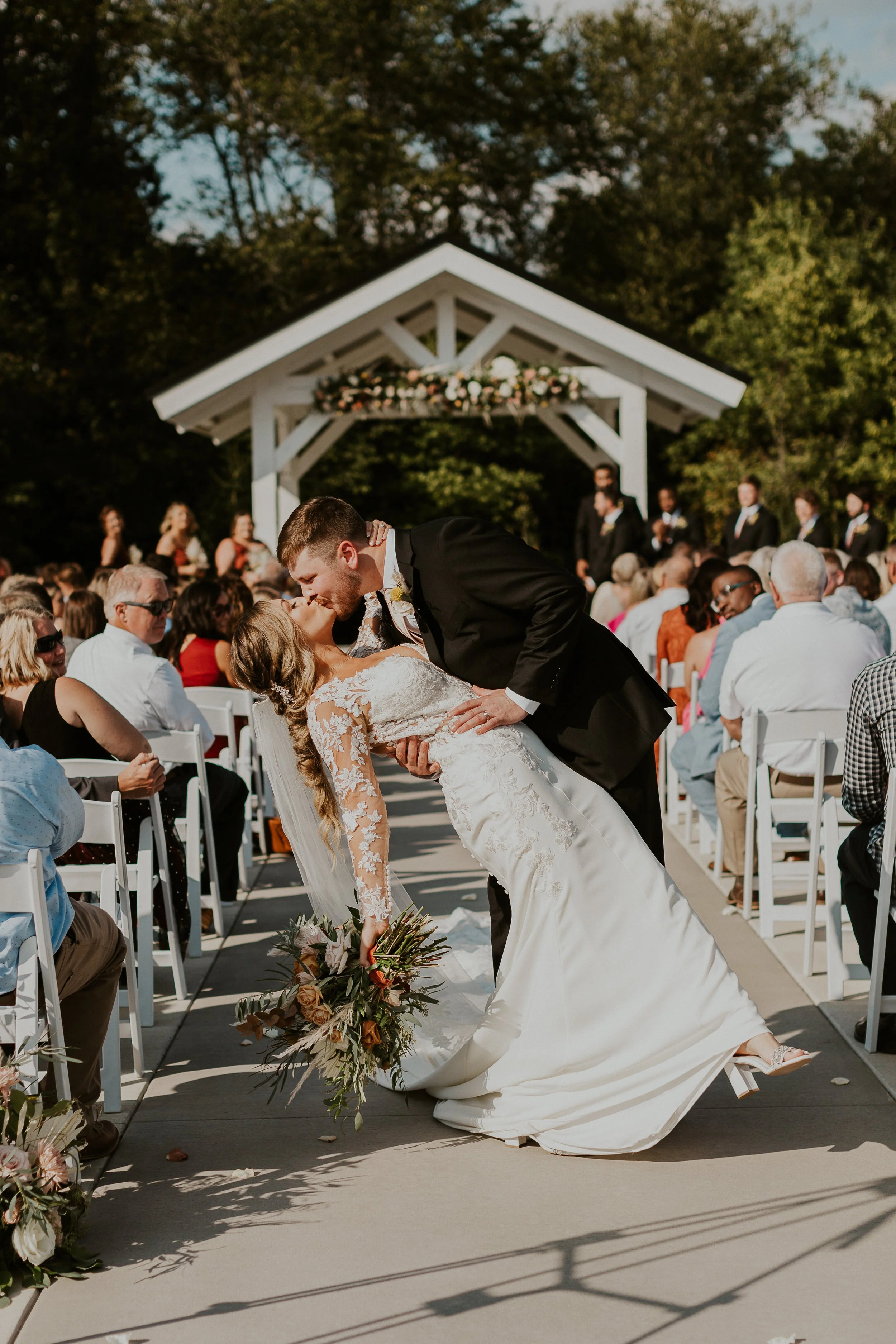 A newlywed couple sharing a kiss during their outdoor wedding ceremony, with the groom dipping the bride, in front of seated guests and a decorated white wedding arch.