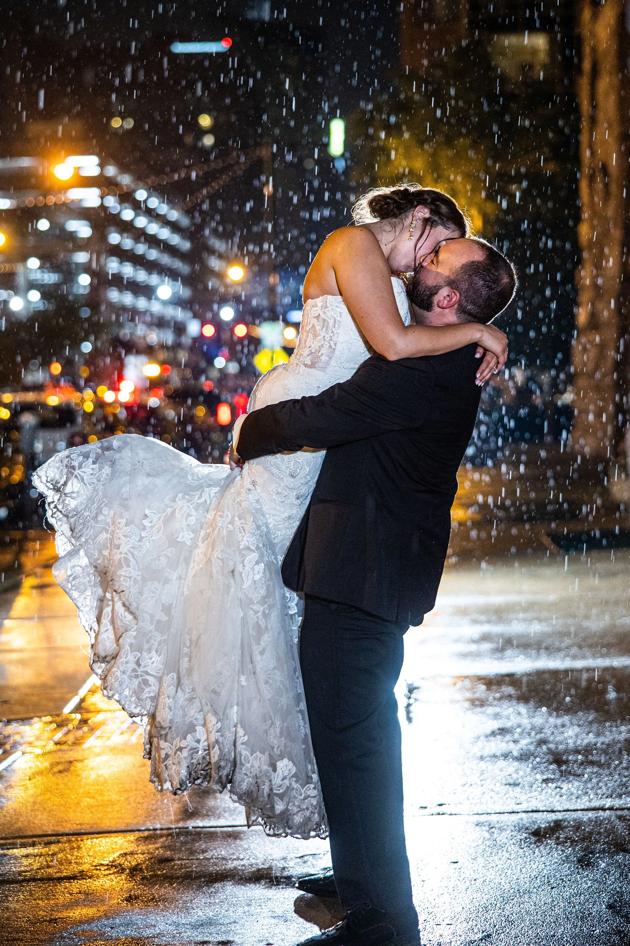 A newlywed couple sharing a kiss outdoors at night in the rain, with city lights in the background. The groom lifts the bride in his arms, her wedding dress flowing.