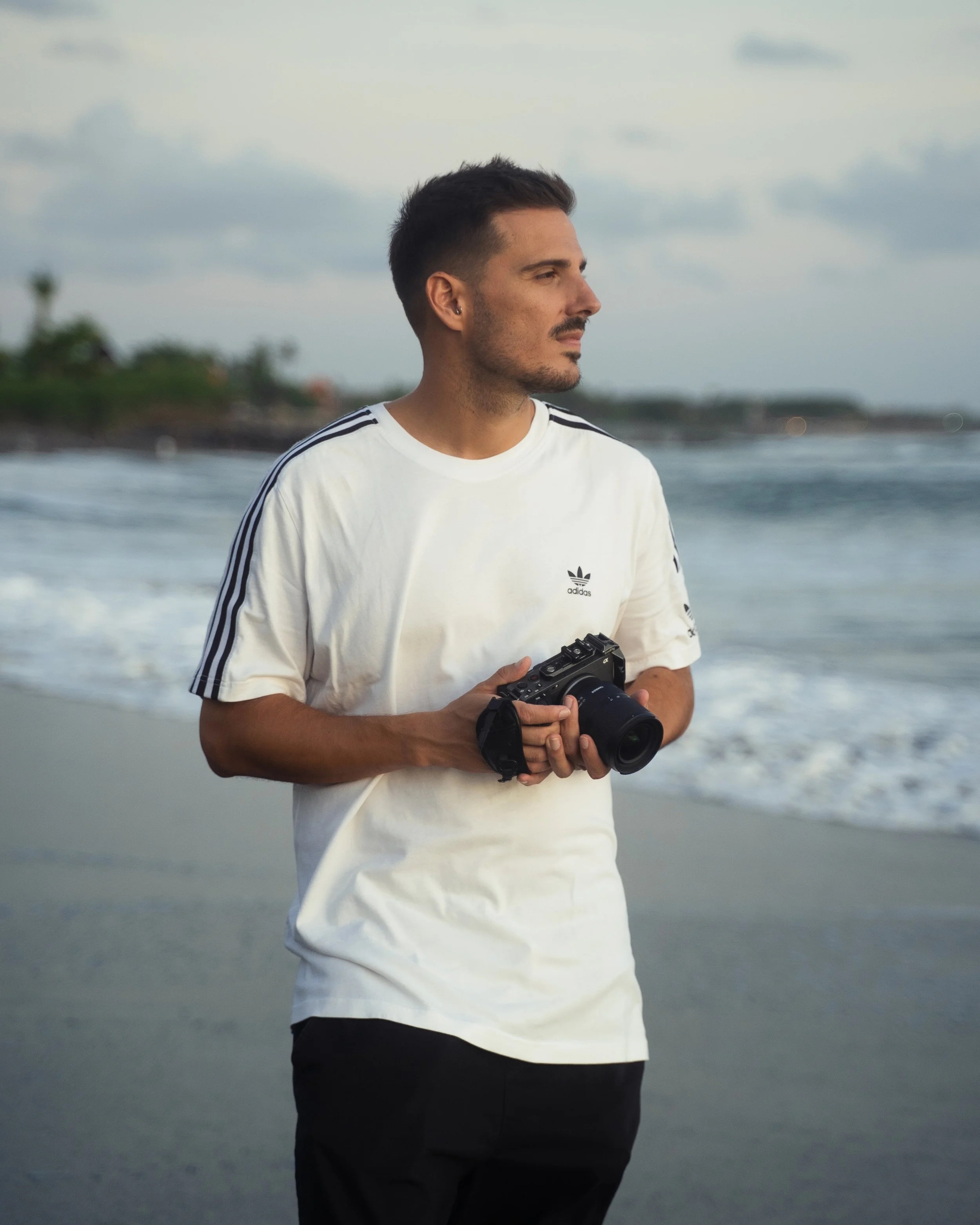Hombre sosteniendo una cámara en la playa, vistiendo una camiseta blanca con rayas en las mangas, mirando hacia el horizonte.