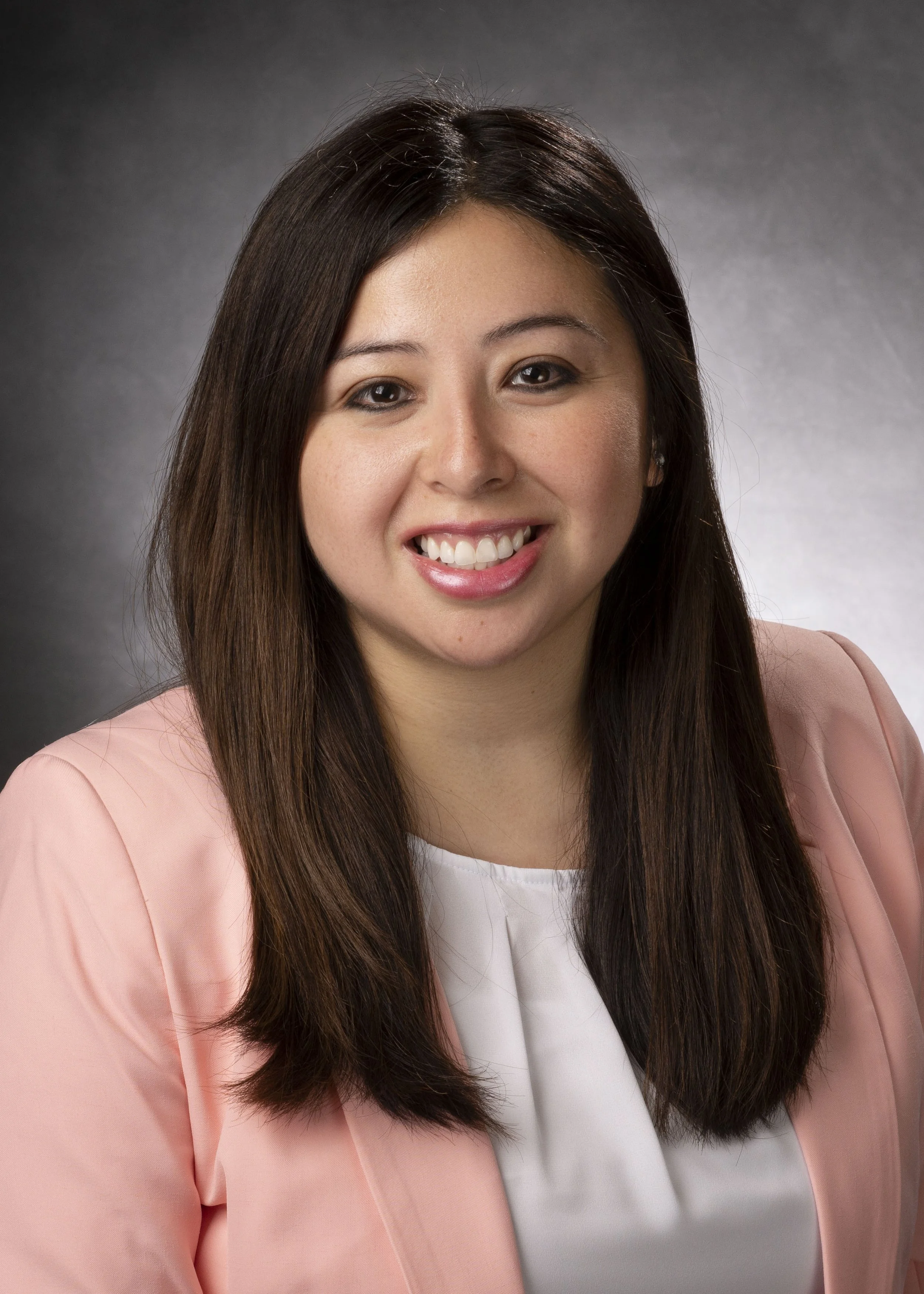 A woman with long, dark brown hair smiling, wearing a light pink blazer and a white top, set against a dark gray background.