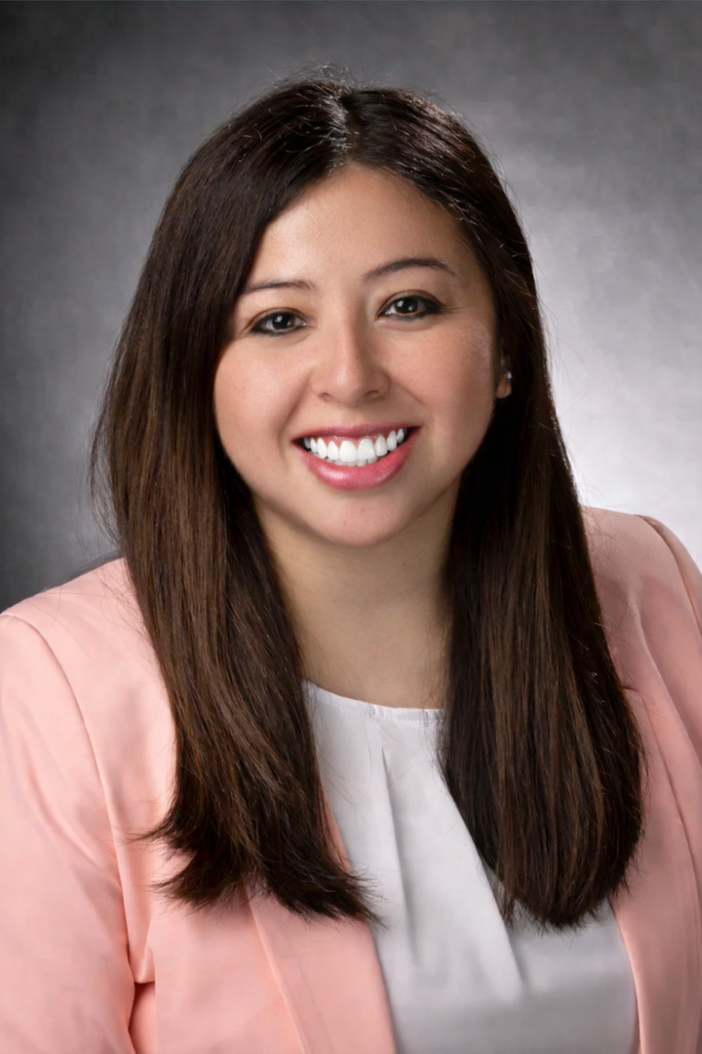 Professional headshot of a woman with long dark hair, smiling, wearing a light pink blazer and white top, against a gray background.