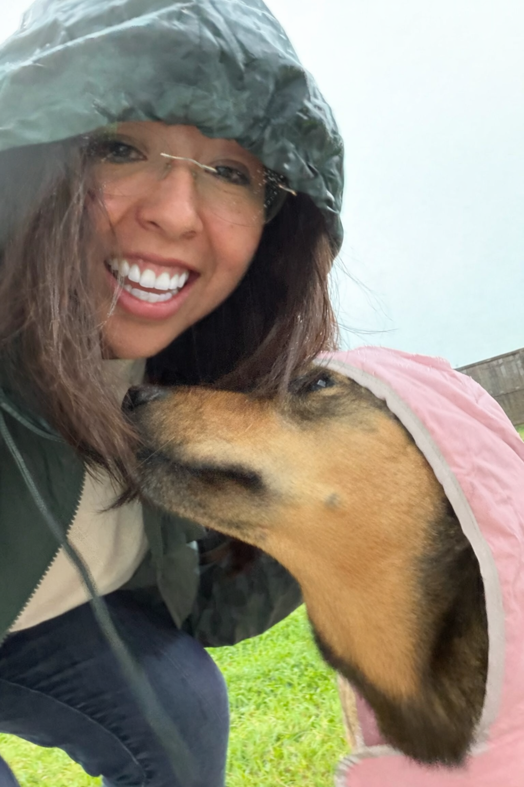 A woman smiling and wearing a rain jacket. She is close to a dog dressed in a pink raincoat, and the dog is licking or nuzzling her face.