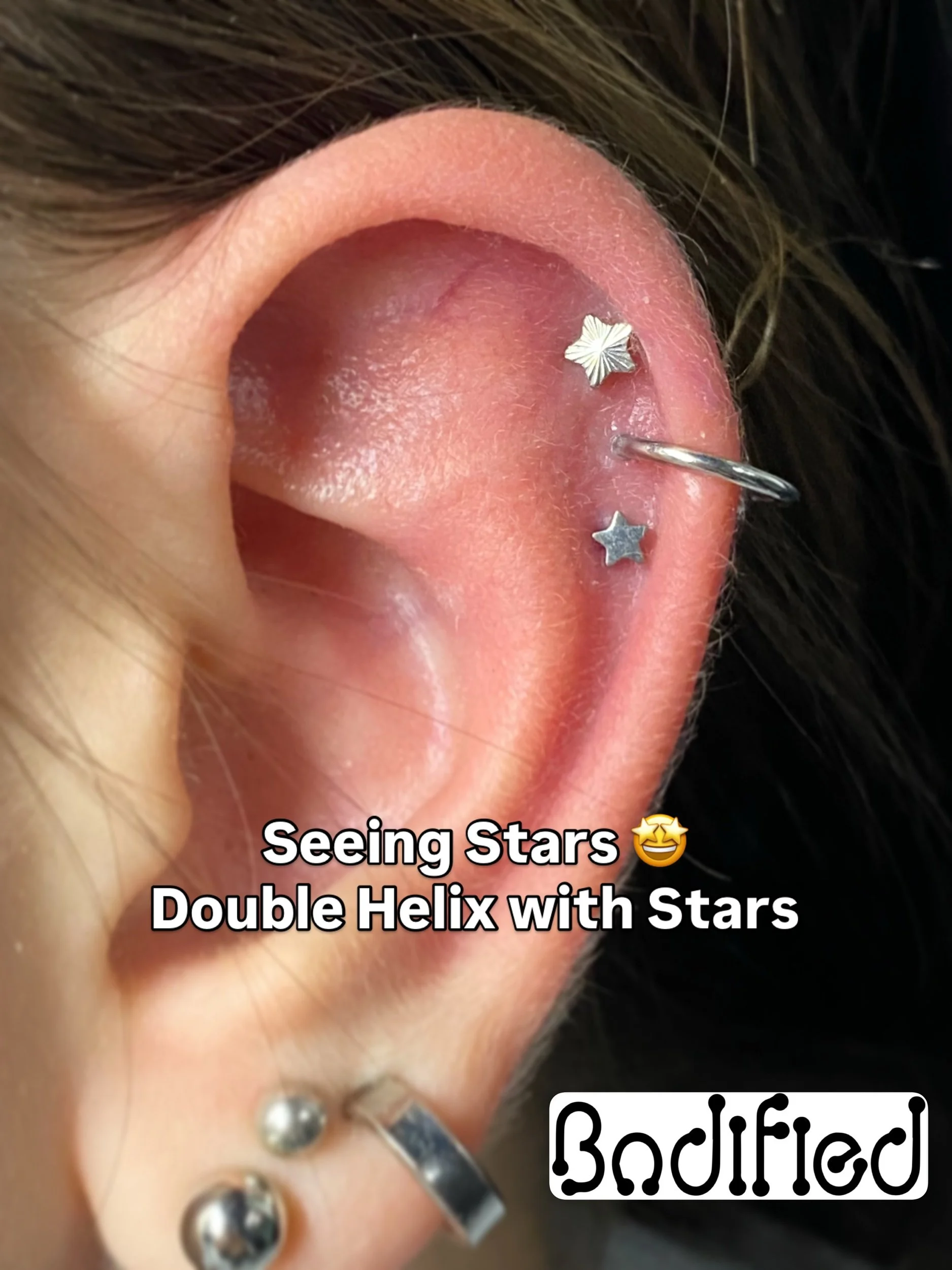Close-up of a person's ear with multiple piercings adorned with star-shaped jewelry, and a caption reading 'Seeing Stars 😄 Double Helix with Stars' and 'Bodified'.