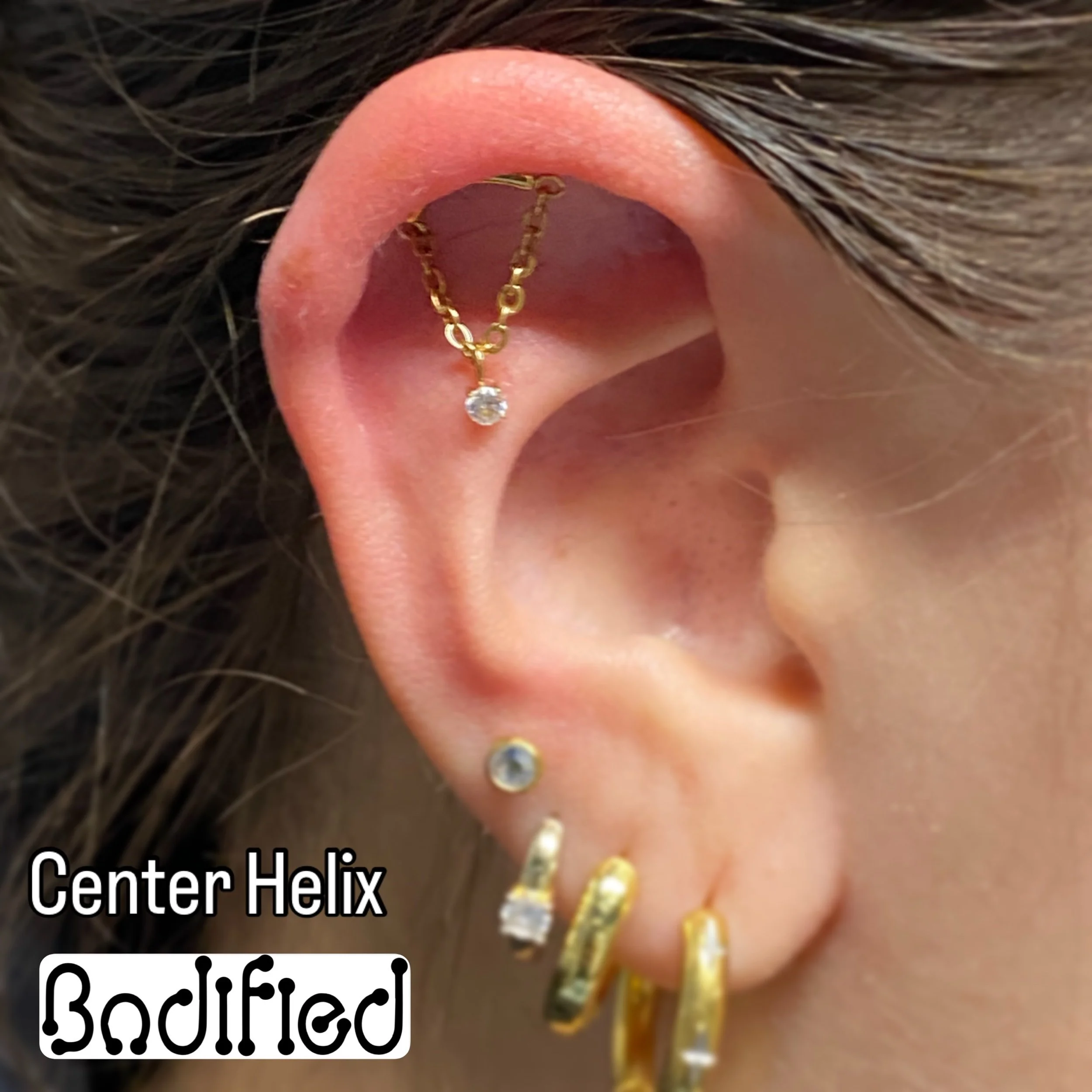 Close-up of a person's ear with multiple gold and gemstone earrings, including a gold chain earring with a dangling clear stone, a small stud with a clear stone, and hoops with clear stones.