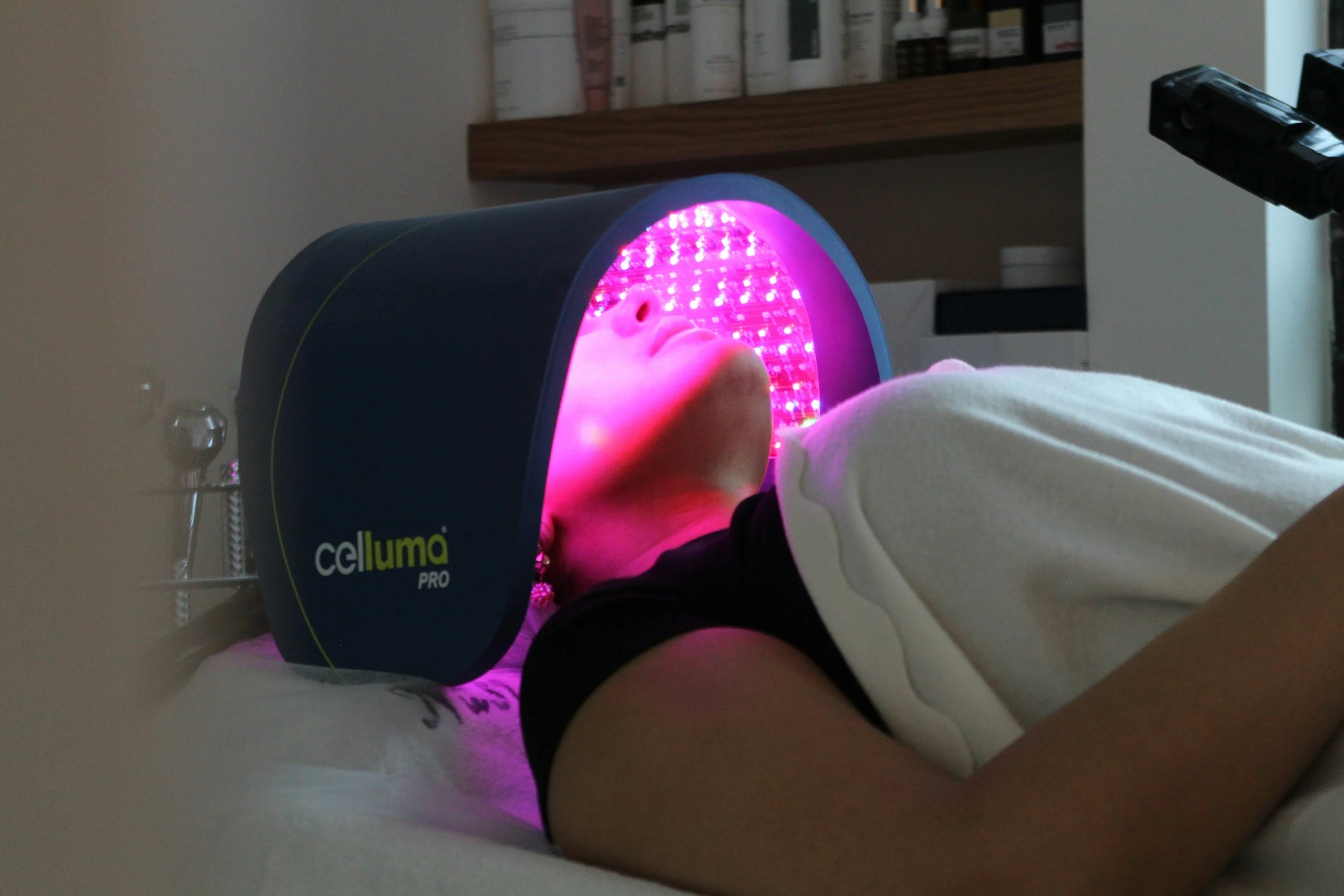 Person lying on a white surface inside a pink and black Celluma Pro LED light therapy device, with their face illuminated by purple light.