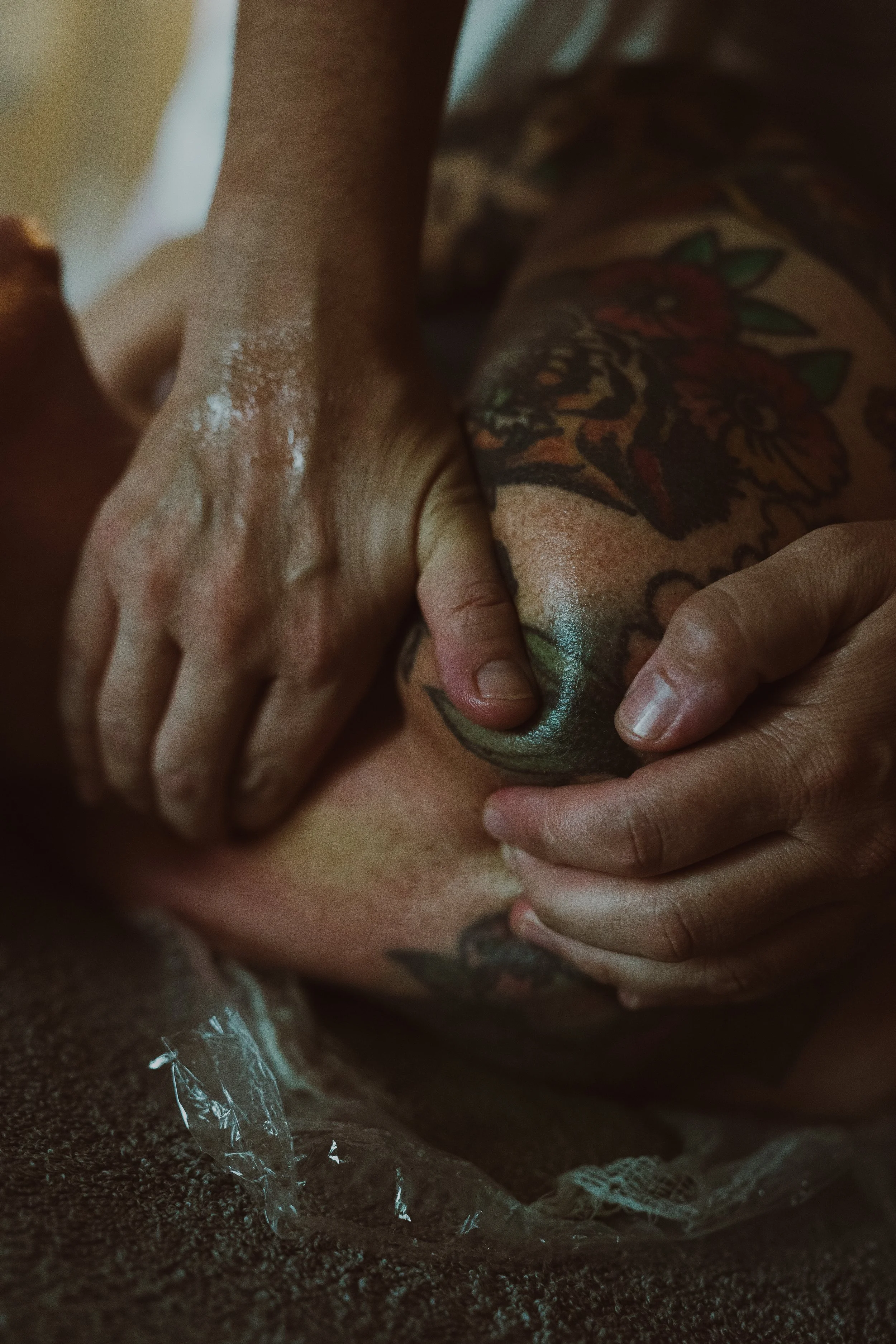 Close-up of a hand pressing a tattooed skin, with another hand near the tattooed area, and a clear plastic bag with small black granular material underneath.