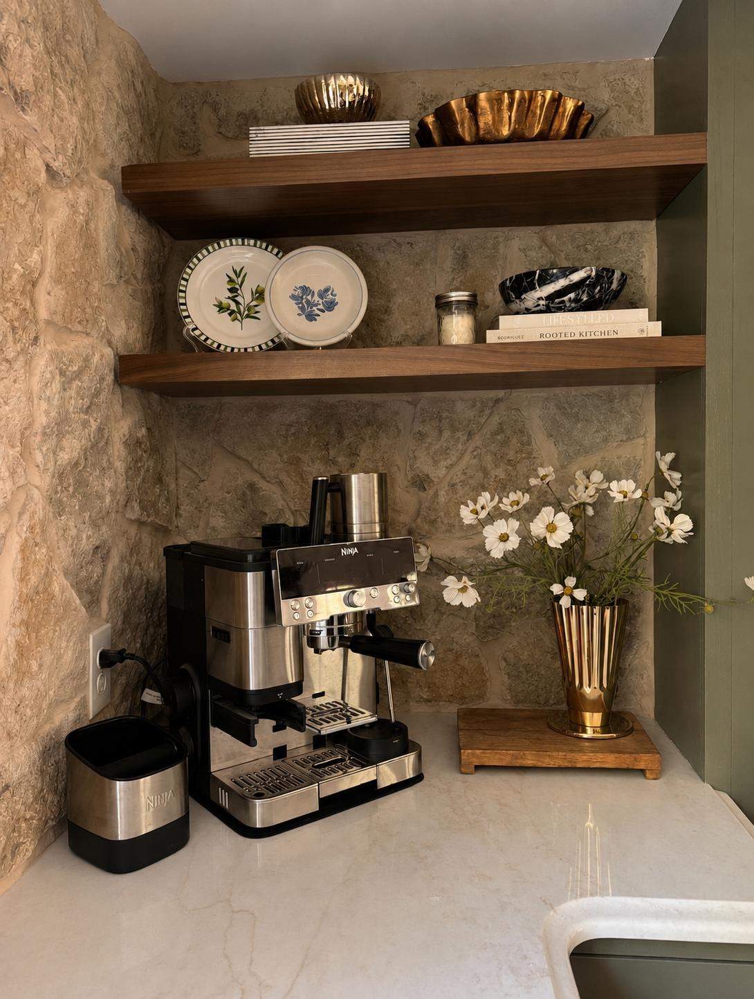 A coffee station with a stainless steel espresso machine, a black and gold vase with white flowers, and decorative plates and bowls on wooden shelves against a stone wall.