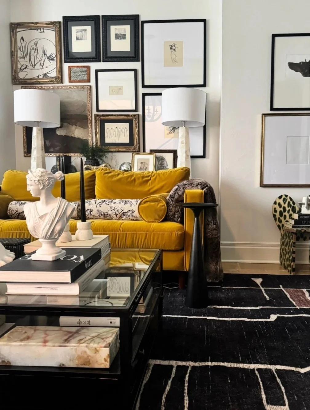 Living room with a yellow velvet sofa, black and white artwork on the walls, white lamps, a glass coffee table with books and sculptures, and a black and beige area rug.