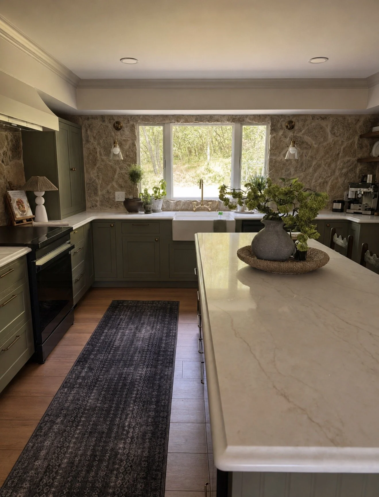 A kitchen with green cabinets, a black stove, a white double sink below a large window, and a marble countertop island with a potted plant. There are various plants on the windowsill and a patterned rug on a hardwood floor.