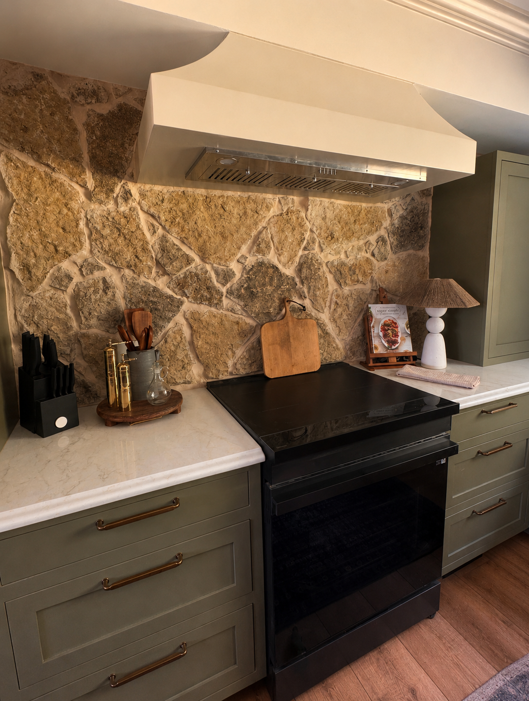 Kitchen with a stone backsplash, black stove, green cabinets with brass handles, and countertop accessories including a cutting board, lamp, and a cookbook on a stand.