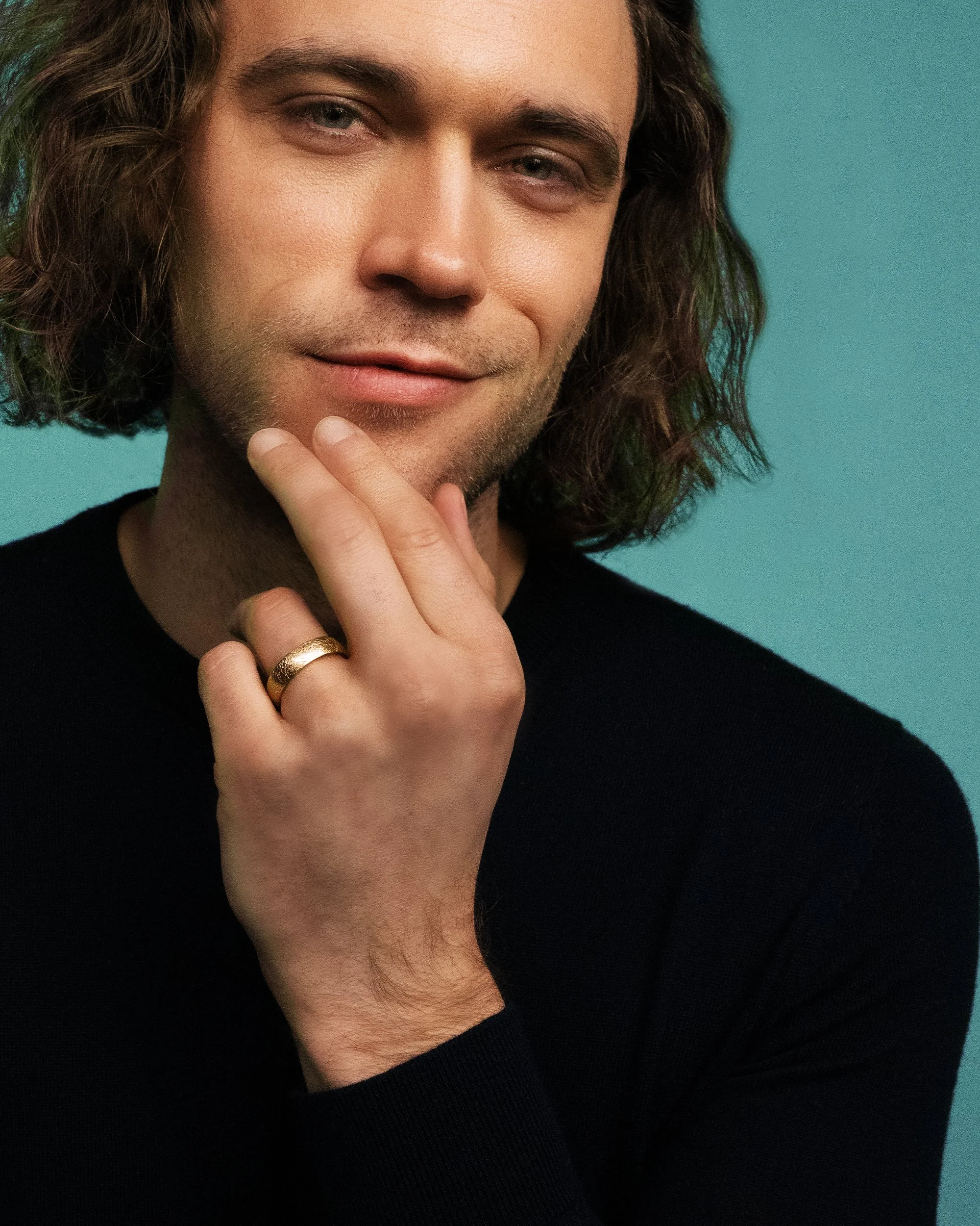 Ein Mann mit schulterlangem Haar trägt einen goldenen Ring am Finger und berührt sein Gesicht, während er leicht lächelt. Hintergrund in Türkis.